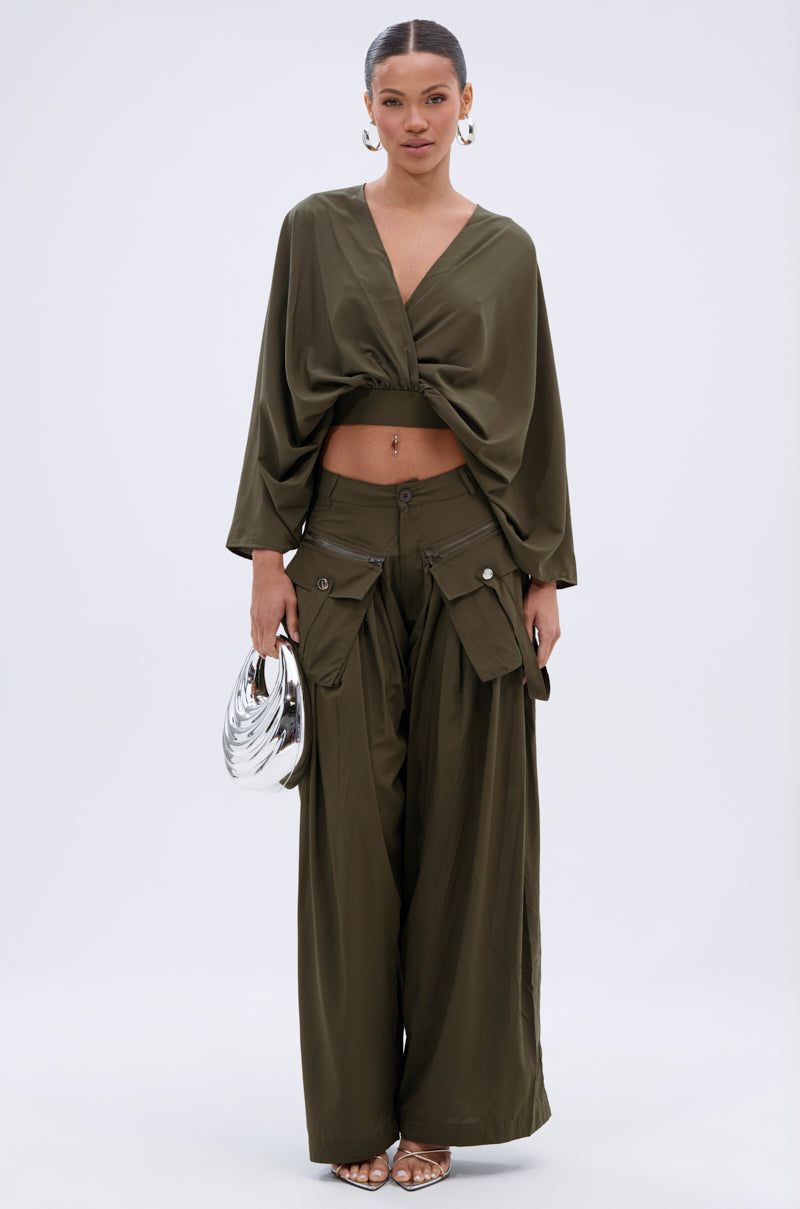 A woman stands against a white background wearing an olive green loose crop top with wide sleeves, the TALK ABOUT ME TROUSER cargo pants, hoop earrings, sandals, and holding a silver metallic handbag. 6