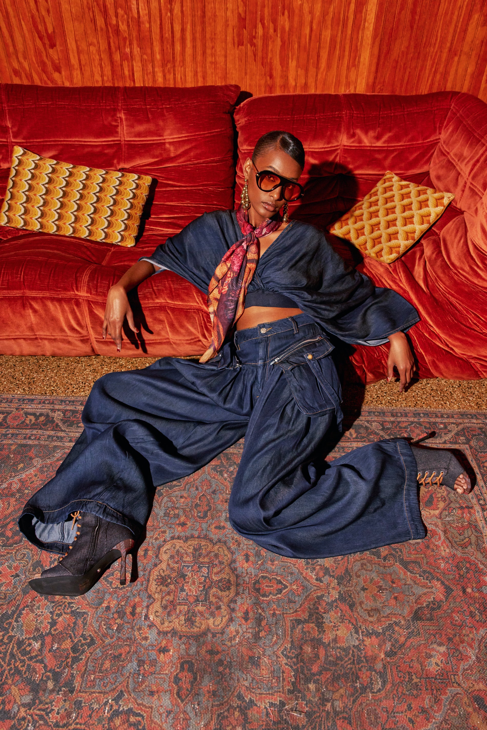 A person wearing the DRIFTING AWAY CARGO DENIM IN DARK BLUE DENIM crop top and wide-leg cargo pants sits on a patterned rug in front of an orange velvet sofa.