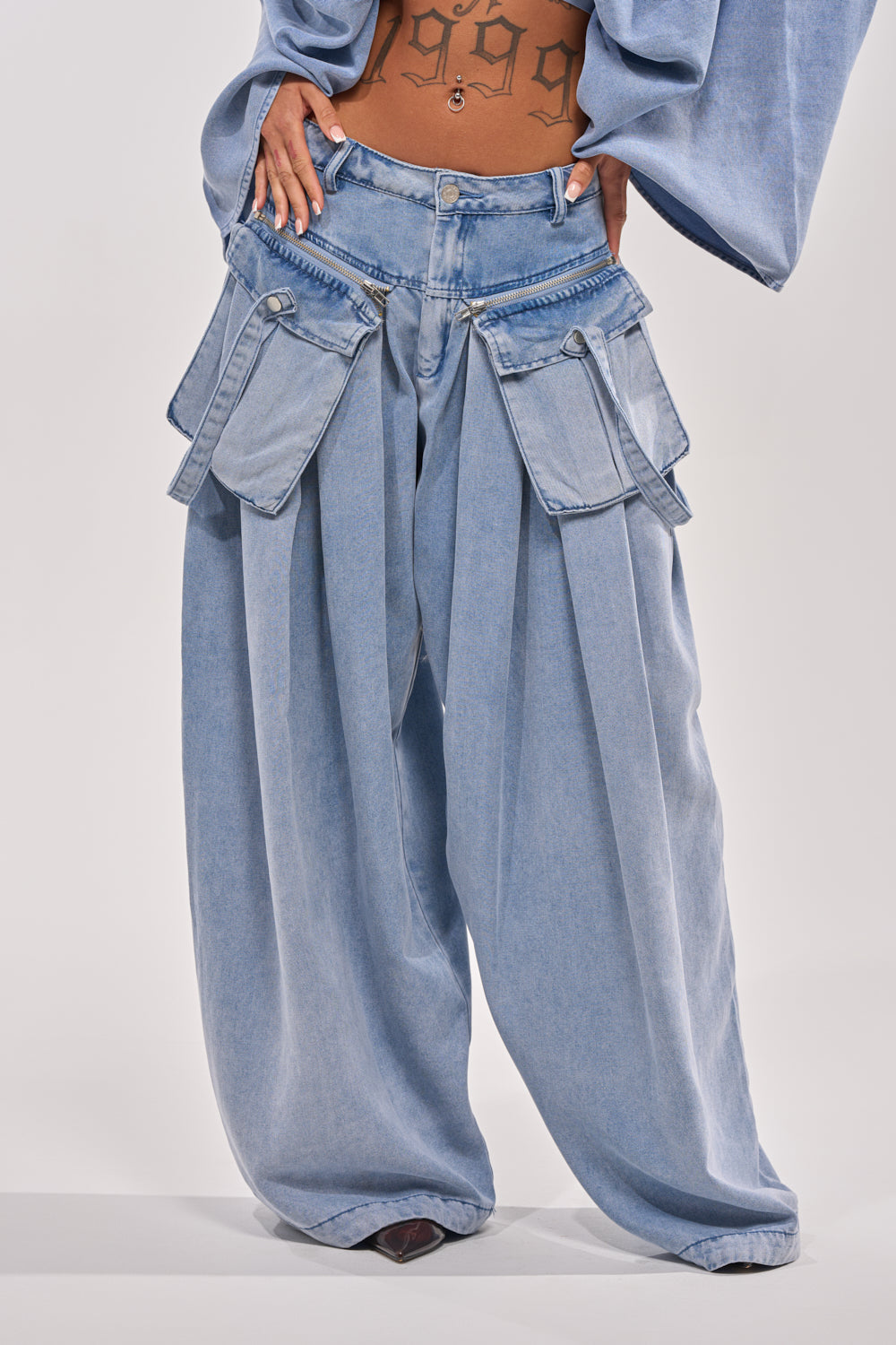 Wearing the DRIFTING AWAY CARGO DENIM—wide-leg pants with large, layered cargo pockets and visible zippers—a bold streetwear style. The midriff and tattooed stomach are visible, face not shown. 1