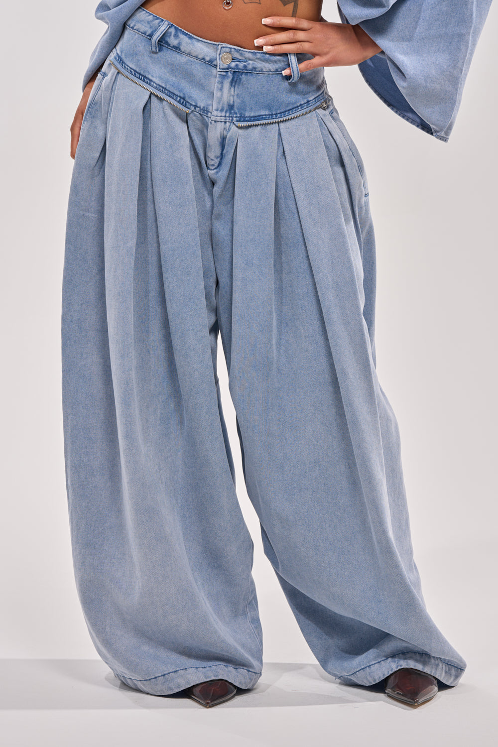 A person models the DRIFTING AWAY CARGO DENIM—high-waisted, wide-leg jeans with pleats and an angled waistband—in a streetwear look, hand on hip. Only their lower body is visible. 2