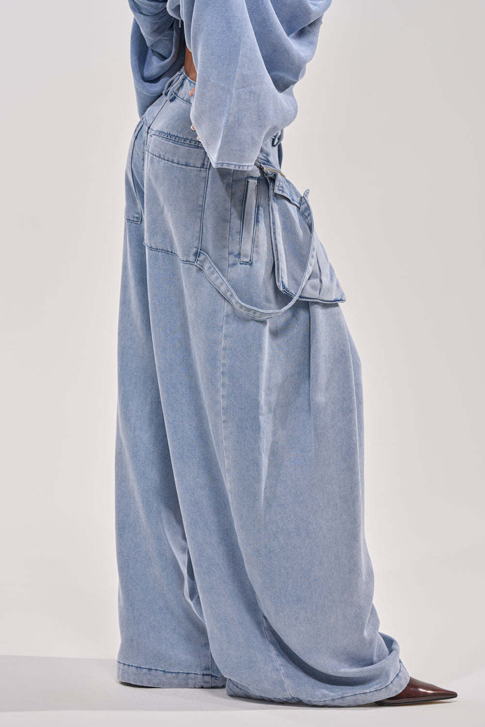 A person in DRIFTING AWAY CARGO DENIM, featuring loose high-waisted pants with large pockets and a matching top, stands sideways on a white background. Only the body from shoulder to feet is shown, conveying a modern streetwear look. 3