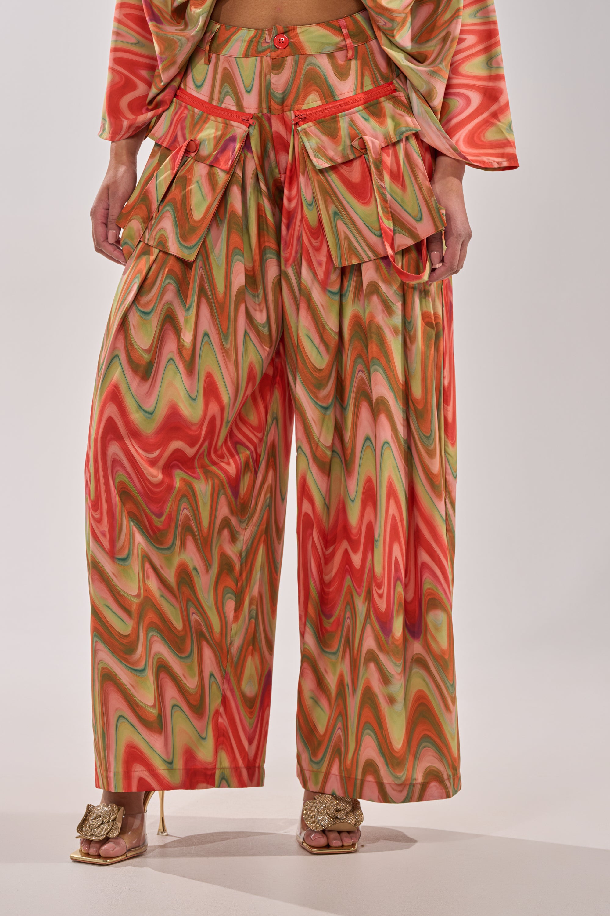 Wearing the DRIFTING AWAY TROUSER—a high-waisted, wide-leg cargo pant with large ruffled pockets—and a matching abstract red, orange, yellow, and green swirl top plus gold open-toe heels for a bold street style look. 1