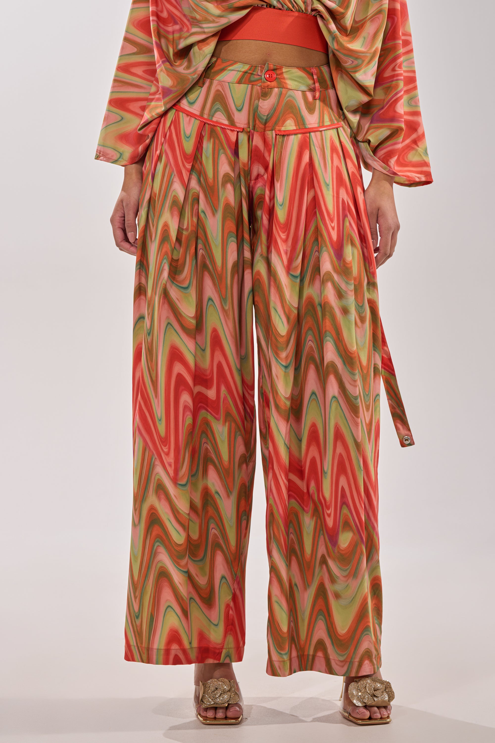 A person showcases statement street style in the DRIFTING AWAY TROUSER—high-waisted, wide-leg cargo pants with a red, orange, and green abstract swirl print—paired with a matching top and heeled sandals. Photo is waist to feet. 4