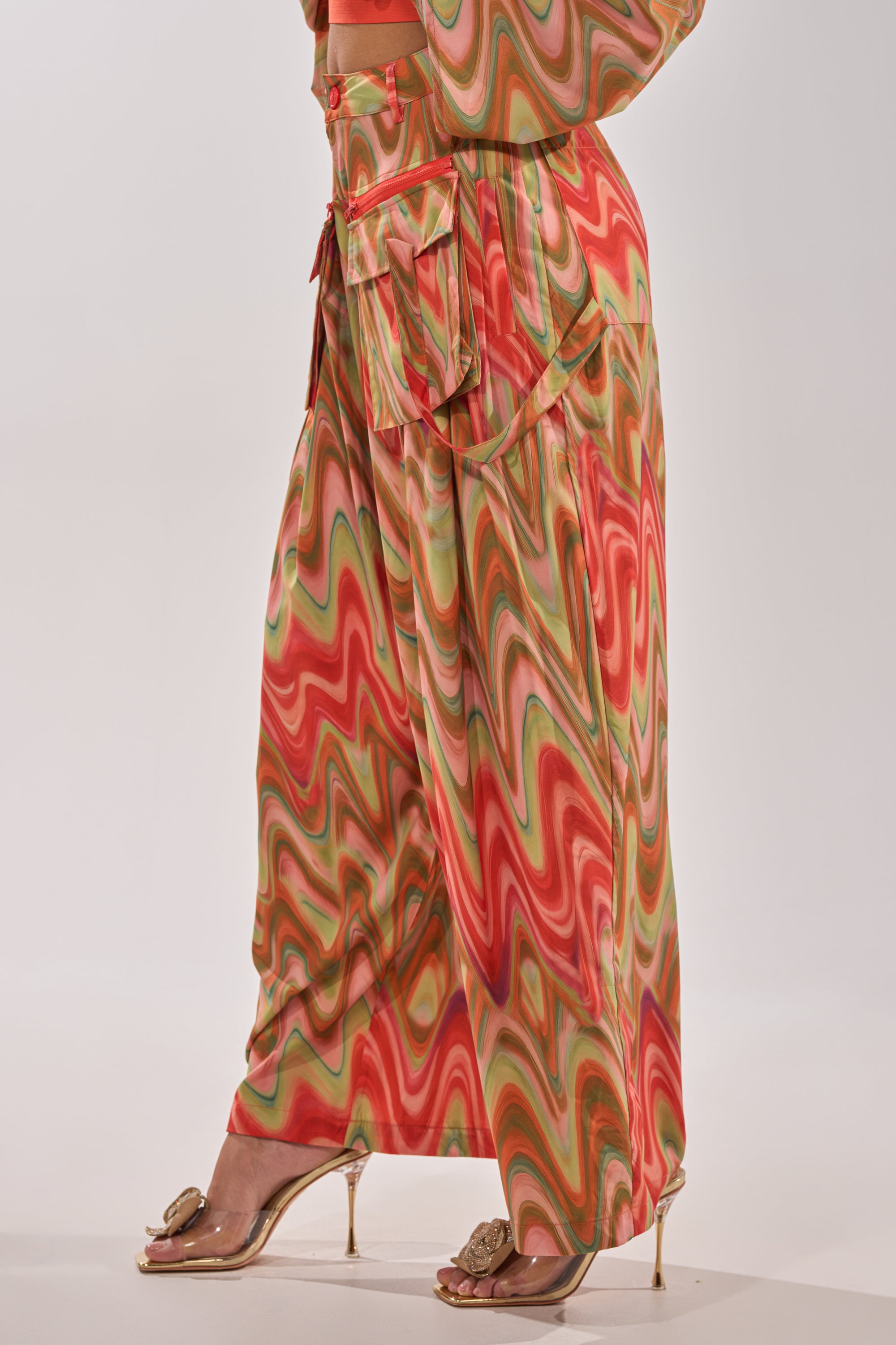 Wearing the DRIFTING AWAY TROUSER—high-waisted, wide-leg cargo pants in an orange, green, and pink abstract swirl print—paired with gold high-heeled sandals for a bold street style look. Only the lower half is visible. 3