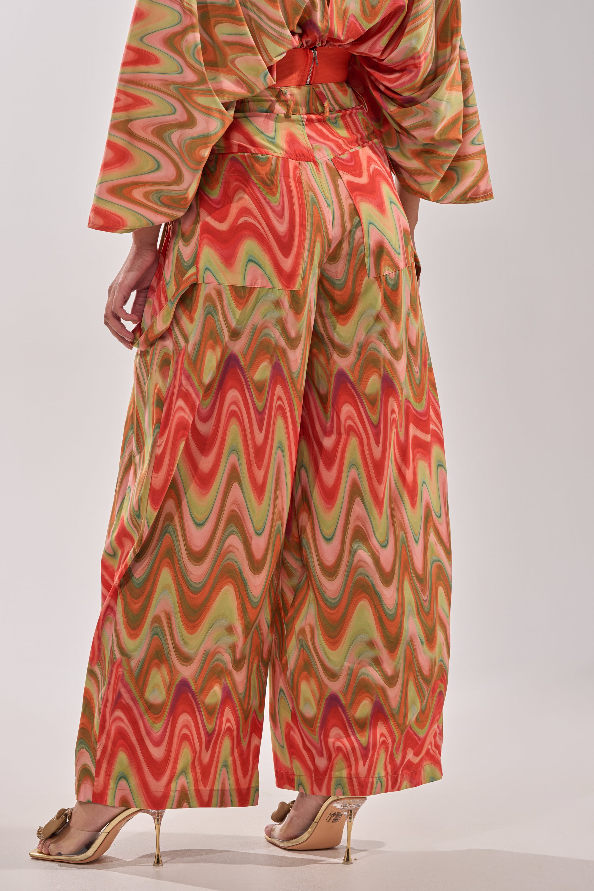 A person models bold street style in the DRIFTING AWAY TROUSER, a high-waisted wide-leg pant with a vibrant abstract swirl print in red, orange, and green, paired with a matching top and high-heeled sandals, facing away from the camera. 5