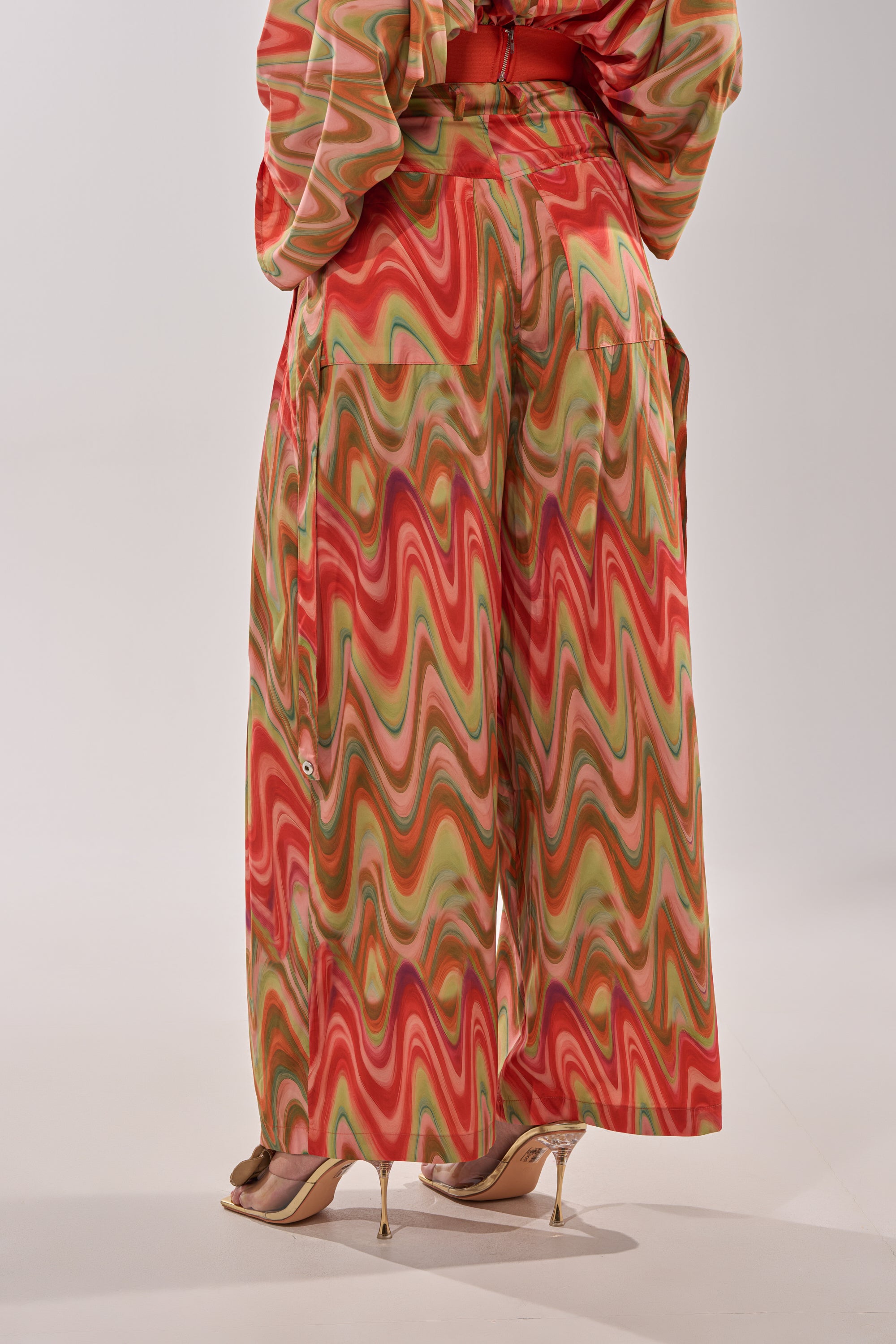 A person stands in heeled sandals wearing the DRIFTING AWAY TROUSER—high-waisted, wide-leg cargo pants with a bold red, green, and pink abstract swirl print. The photo, shot from behind, highlights their statement street style. 6