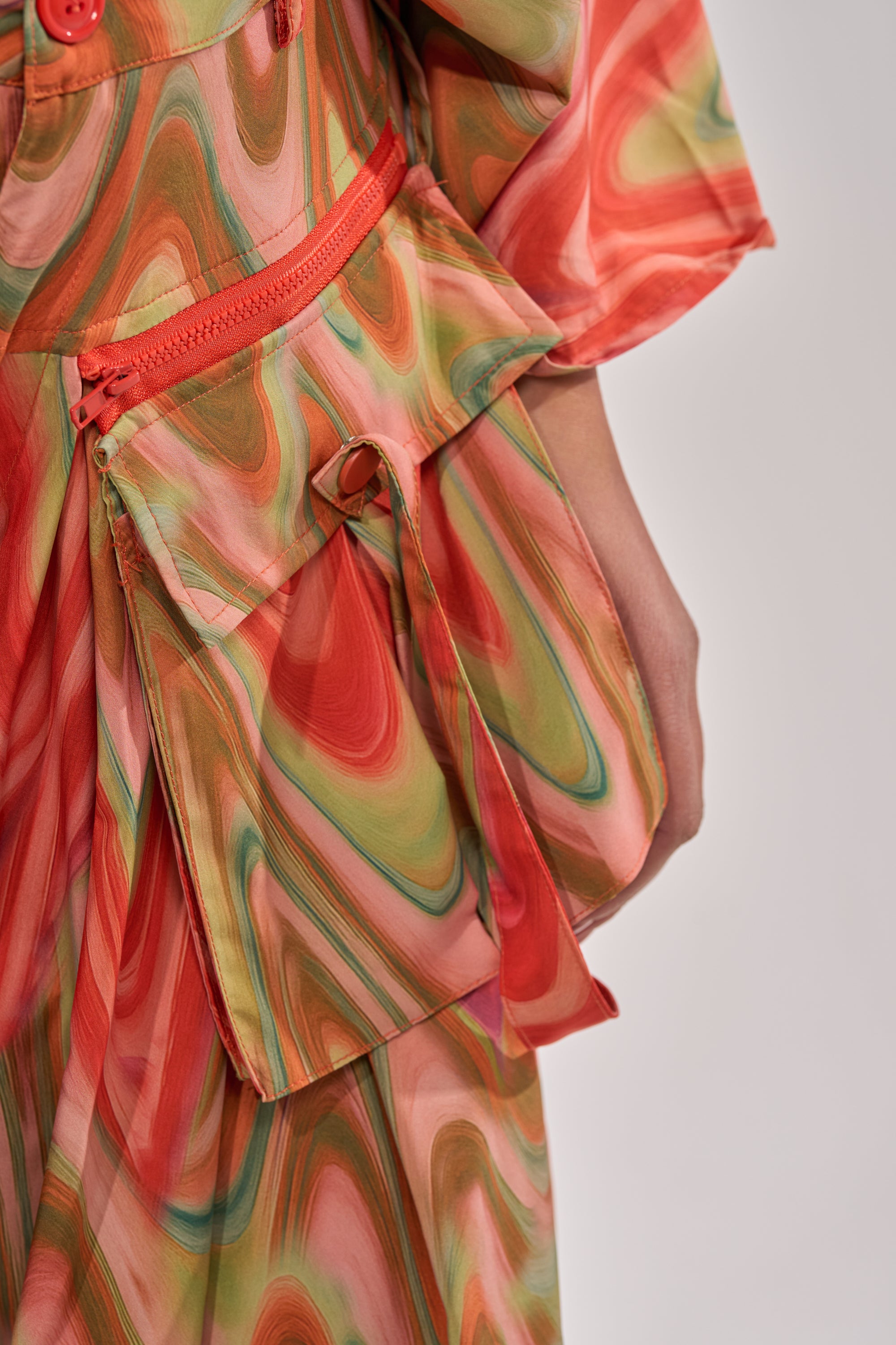 Close-up of the DRIFTING AWAY TROUSER—high-waisted, wide-leg cargo pants featuring an abstract red, green, and pink swirl print. Zipper pocket and button details highlight the flowing fabric for bold street style. A hand rests beside the pocket. 8
