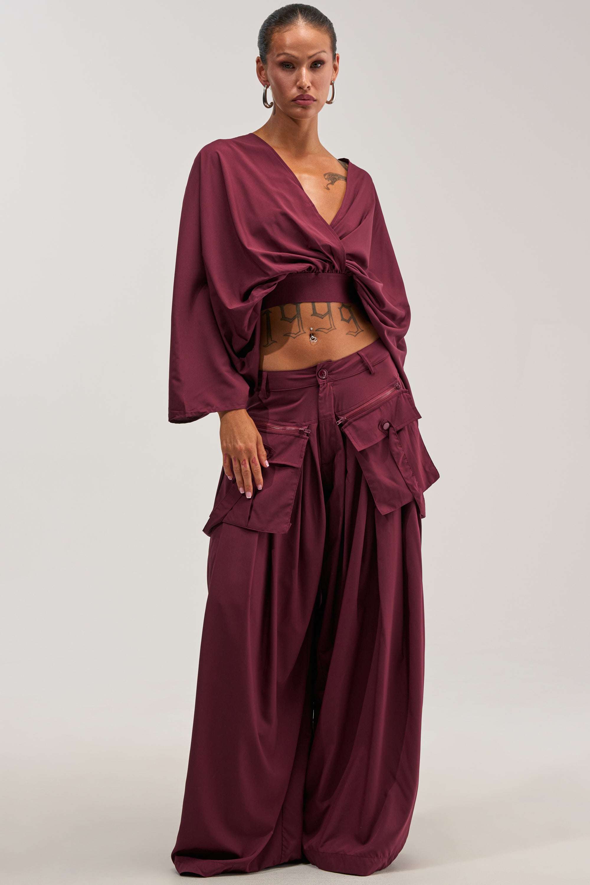 A woman stands against a plain light background, wearing loose-fitting streetwear that includes the TALK ABOUT ME TROUSER IN DEEP BURGUNDY—wide-leg cargo pants with large pockets—paired with a matching deep burgundy wrap top.