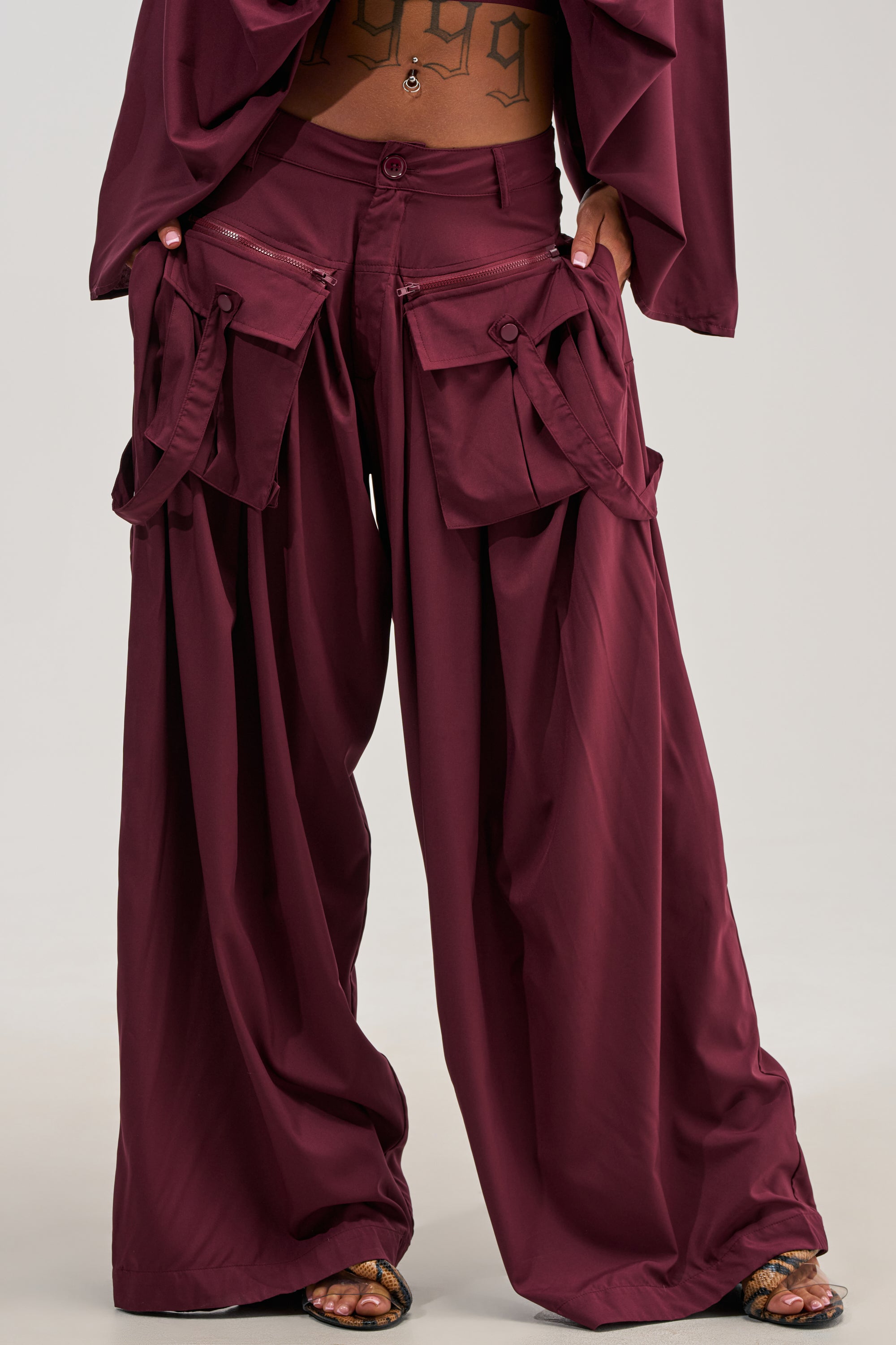A person wearing the TALK ABOUT ME TROUSER IN DEEP BURGUNDY—loose, wide-leg cargo pants with large front pockets—paired with a matching top and sandals. Their hands are tucked into the streetwear trousers’ pockets, revealing their midriff and a visible tattoo.