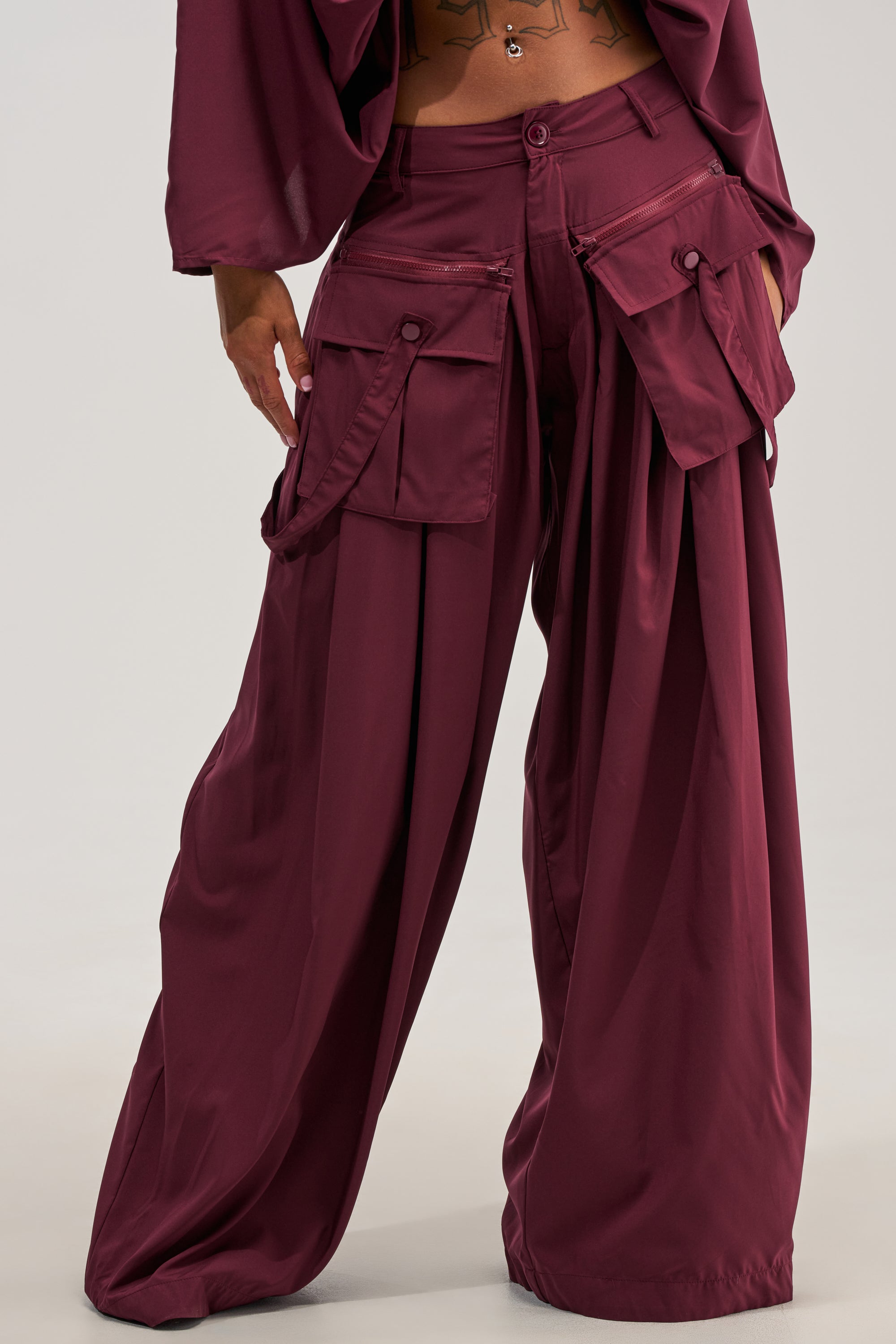 A person is shown wearing the TALK ABOUT ME TROUSER IN DEEP BURGUNDY, featuring a wide-leg silhouette, large flap pockets, and braided detailing. Only the lower torso and upper legs are visible against a plain, light-colored background.