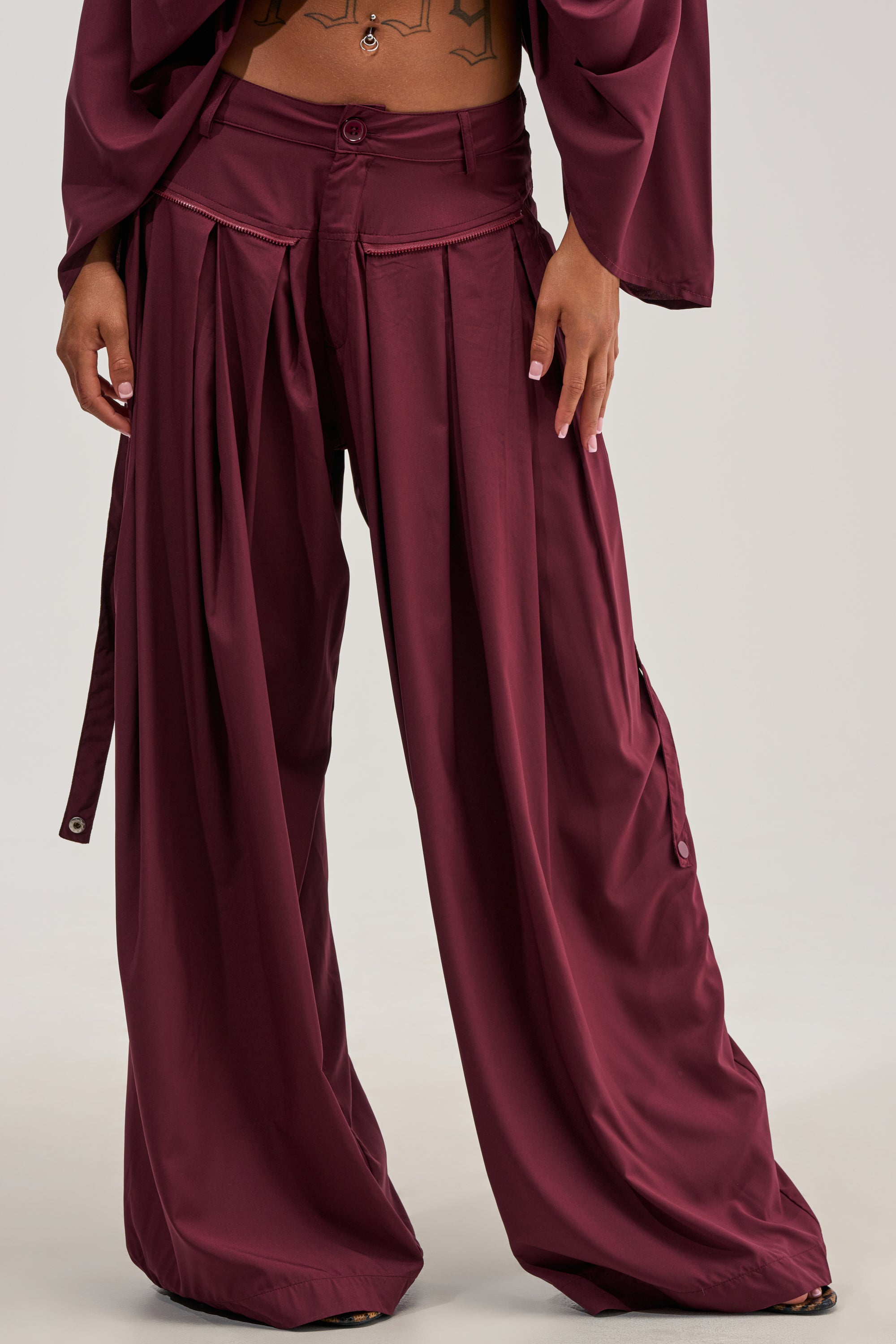 A person is wearing the TALK ABOUT ME TROUSER IN DEEP BURGUNDY, which are loose-fitting and high-waisted with pleats, paired with a matching top. Their midriff and tattooed torso are visible, and their hands rest at their sides.