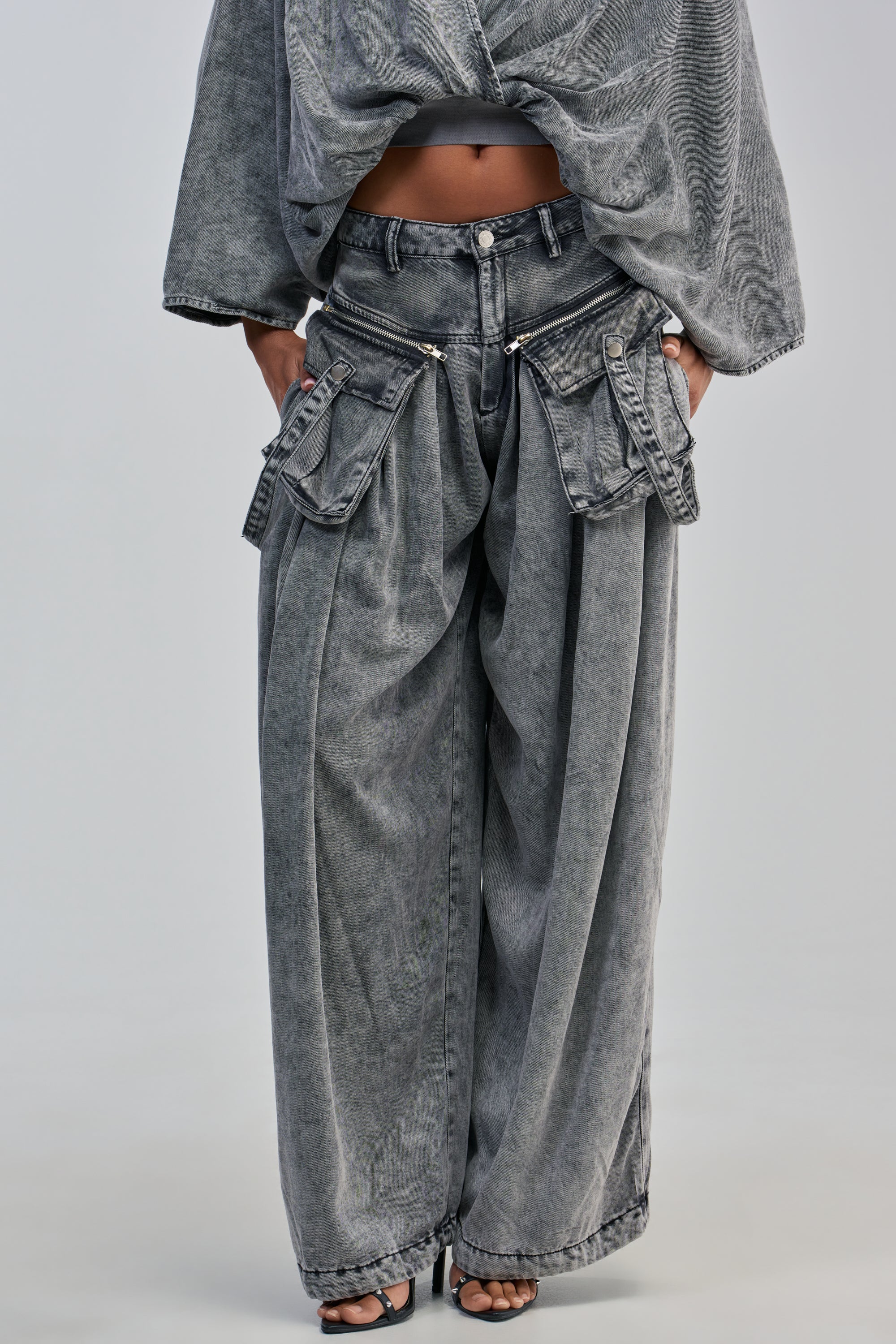 Person wearing the DRIFTING AWAY CARGO DENIM IN CHARCOAL, featuring a wide-leg silhouette, acid-wash gray finish, large zipper pockets, and utility styling for a modern, oversized look. Only the lower half of the body is visible.