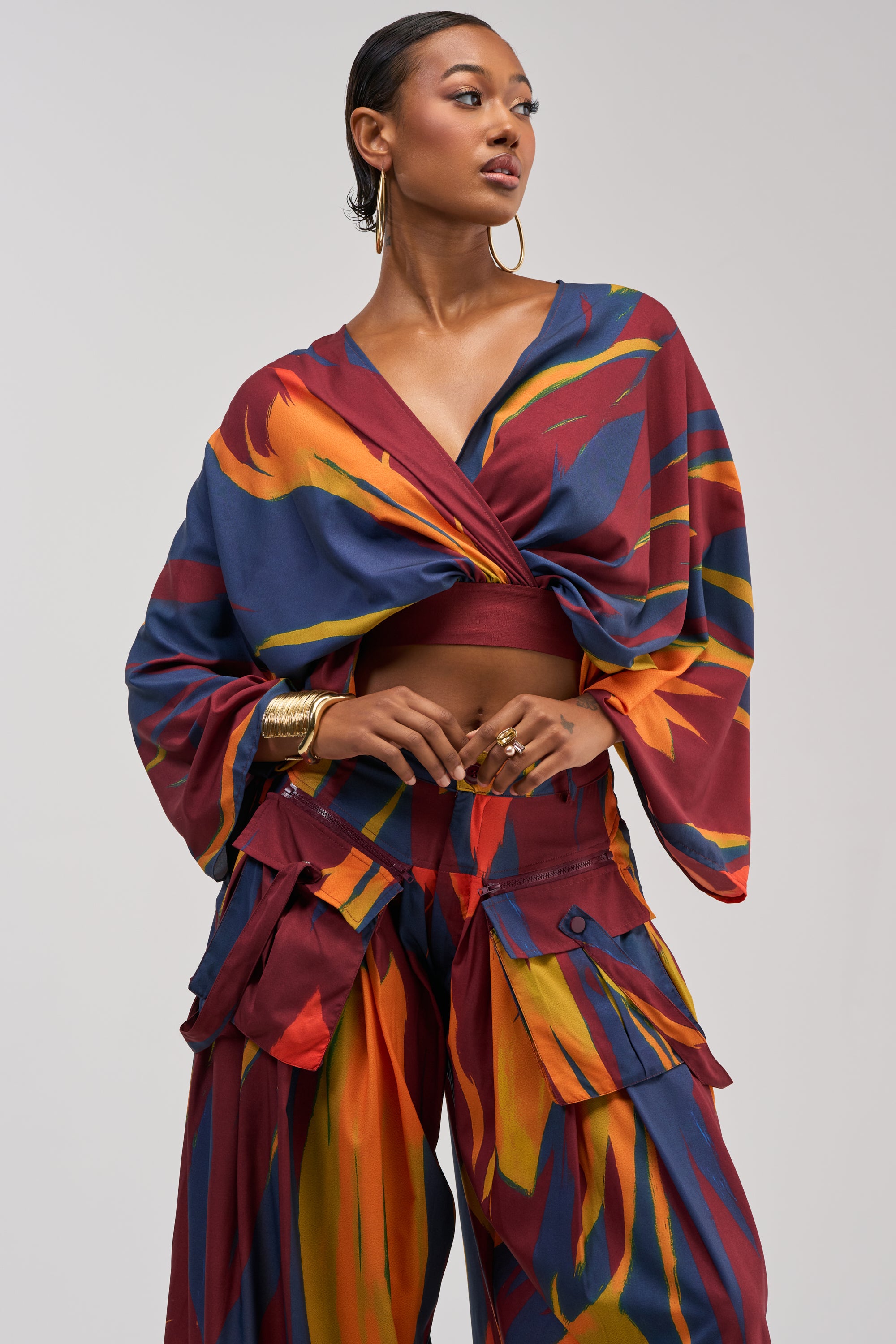 A woman models the TAKE ME AWAY BLOUSE, featuring a vibrant, abstract print and wide sleeves. She pairs the cross-front, cropped blouse with loose pants and accessorizes with gold hoop earrings and bangles, creating a modern, art-inspired look against a plain background.