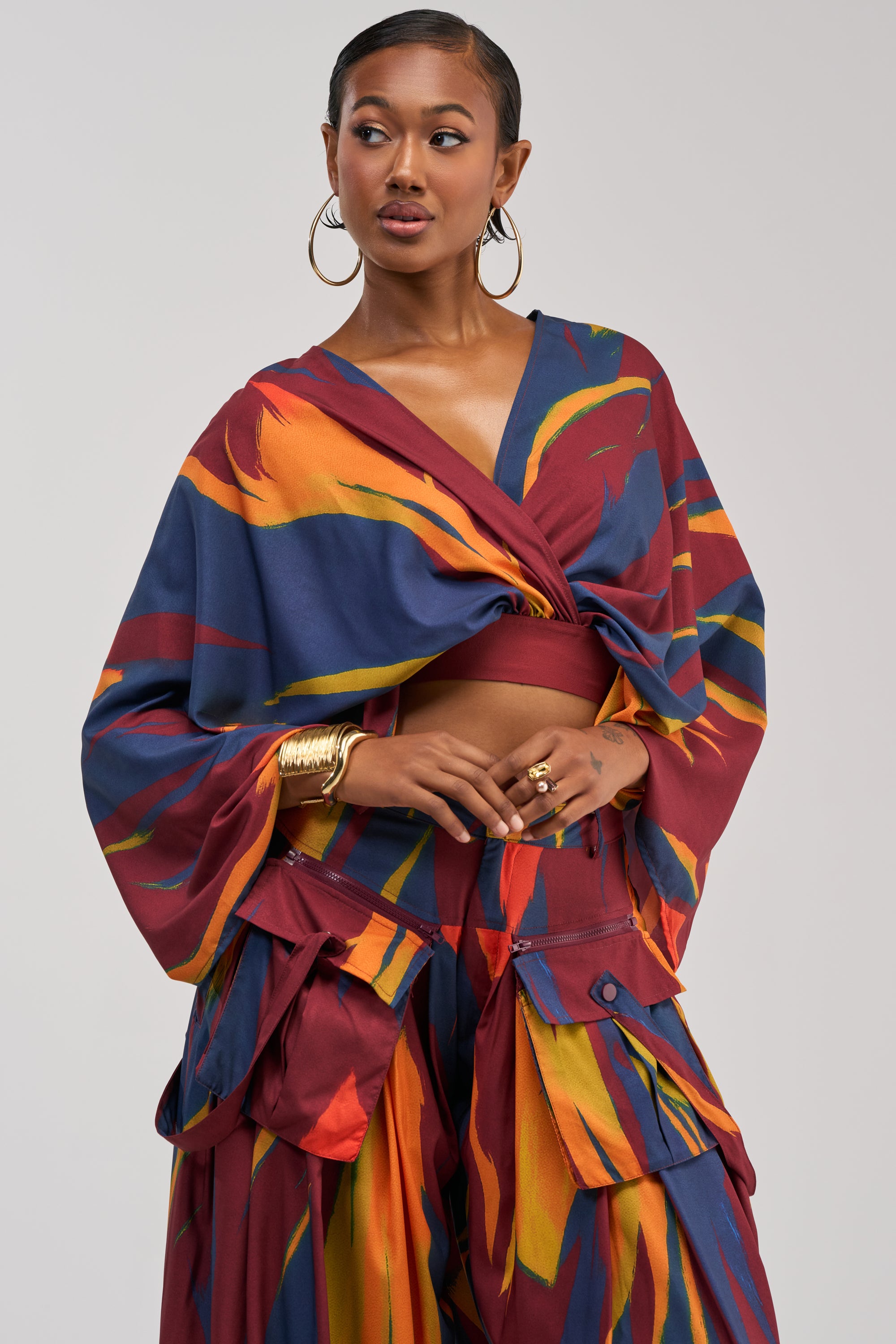 A woman wears the TAKE ME AWAY BLOUSE, a vibrant, multicolored cross-front cropped top with bold red, blue, orange, and yellow patterns. She pairs it with wide-leg pants and accessorizes with gold hoop earrings and bracelets, her hair slicked back.