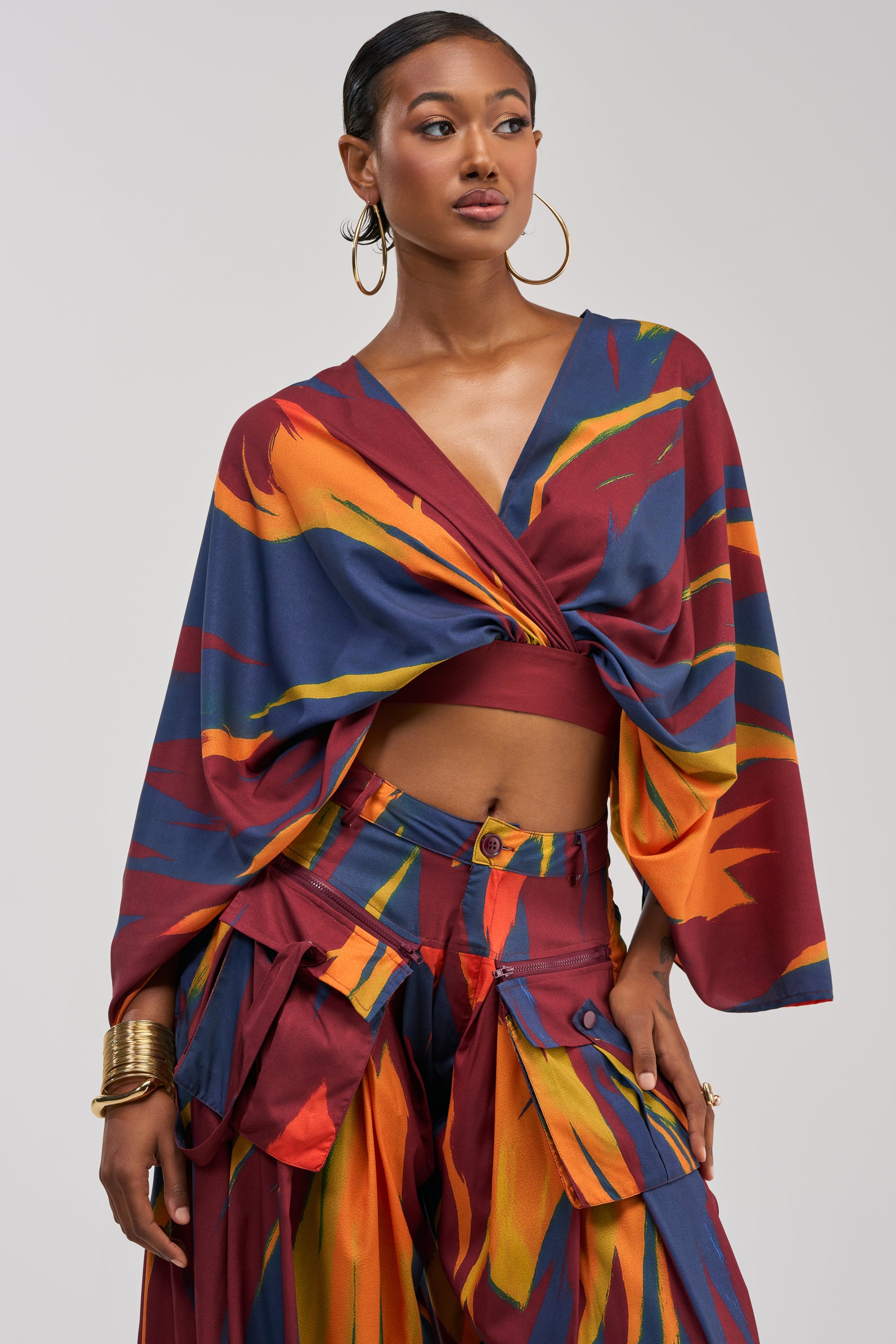 A woman stands confidently with one hand on her hip against a plain background, wearing the TAKE ME AWAY BLOUSE—a vibrant, abstract print top with wide sleeves—paired with large hoop earrings and gold bangles.