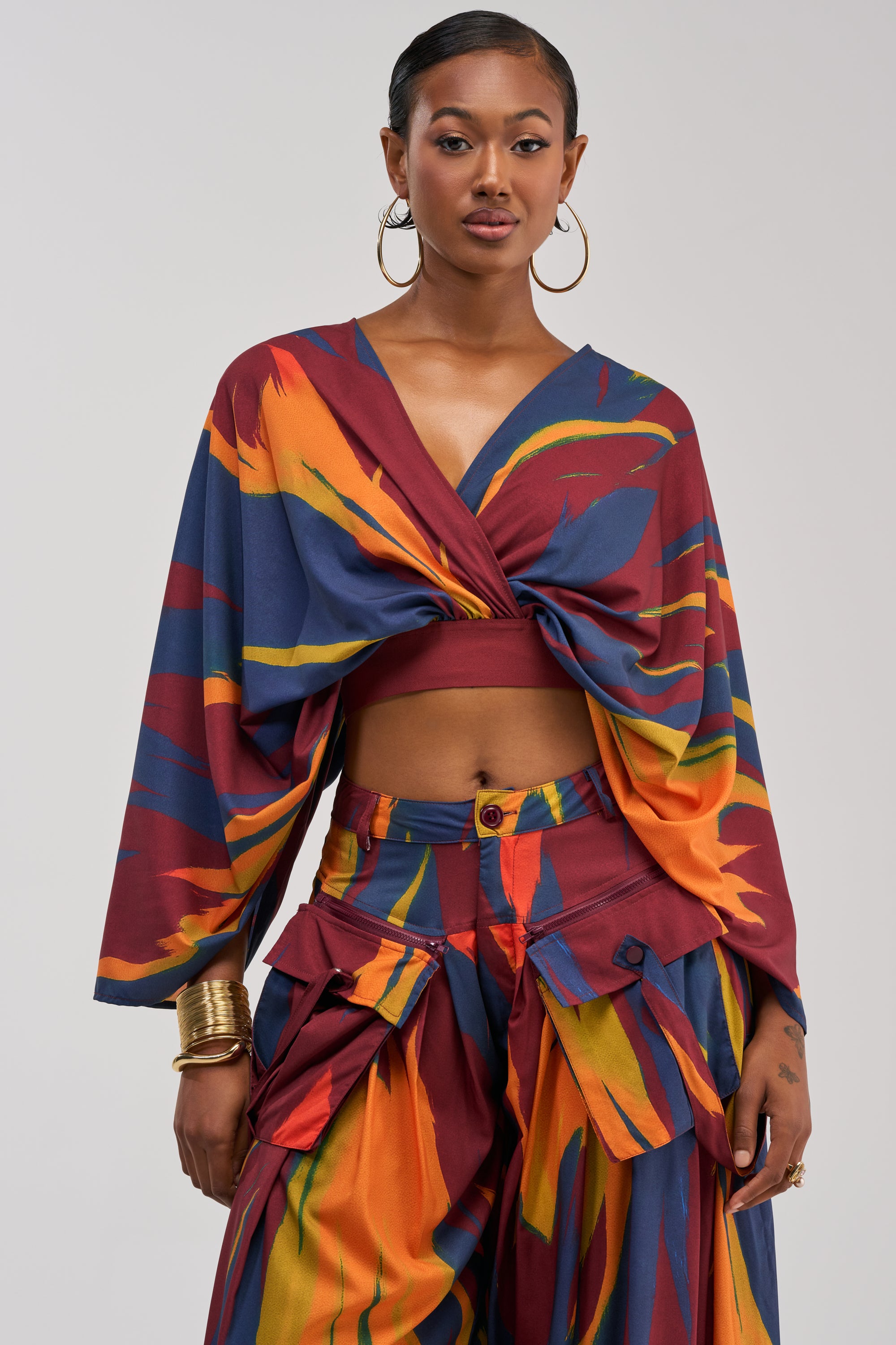 A woman wearing the TAKE ME AWAY BLOUSE, styled as a knotted crop top with wide sleeves and an abstract, colorful print, paired with high-waisted pants featuring large pockets, posed against a plain background.