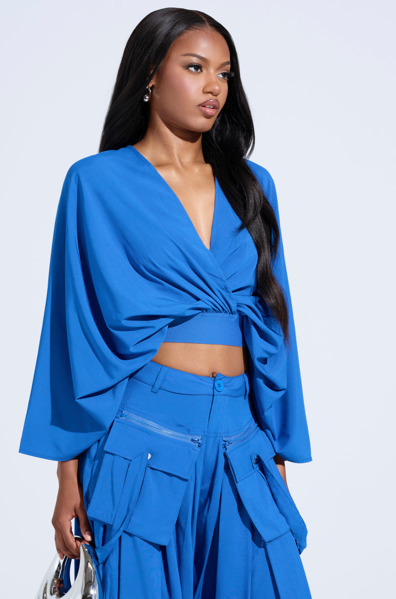 Woman in a vibrant Talk About Me Blue Blouse with flowy dolman sleeves, paired with a long-sleeve knotted crop top, cargo pants, and holding a shiny silver handbag against a light background.