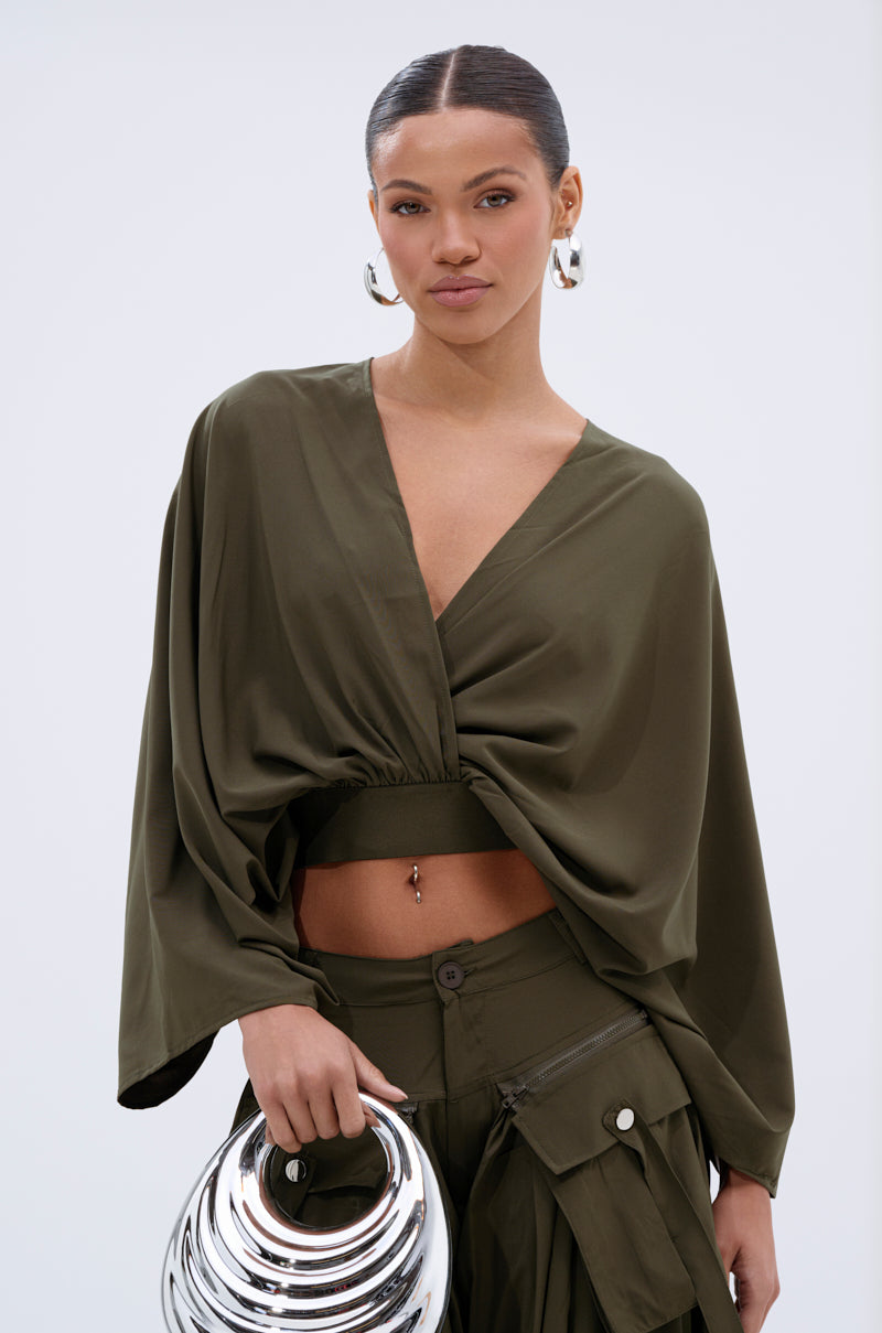 A woman with slicked-back hair wears the TALK ABOUT ME BLOUSE in olive green, styled with wide batwing sleeves and a surplice neckline, paired with matching cargo pants and a silver circular handbag, against a plain light background.