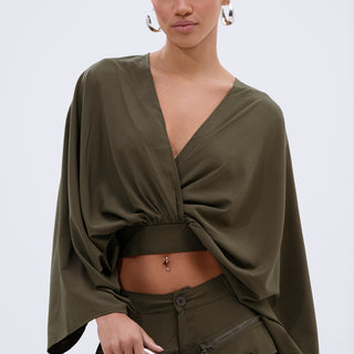 A woman with slicked-back hair wears the TALK ABOUT ME BLOUSE in olive green, styled with wide batwing sleeves and a surplice neckline, paired with matching cargo pants and a silver circular handbag, against a plain light background.