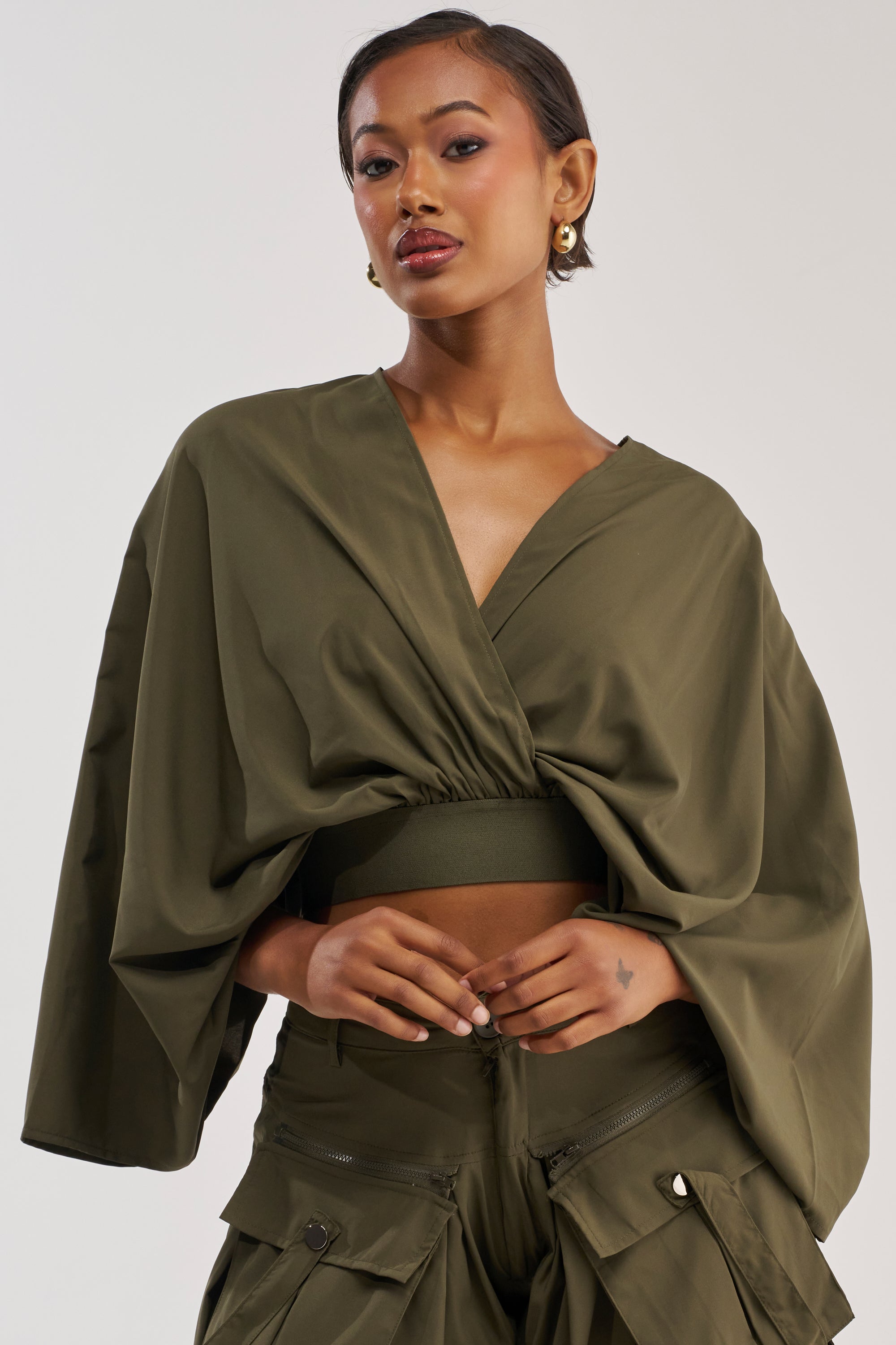 A woman wears the TALK ABOUT ME BLOUSE, an olive green kimono-style top with batwing sleeves, paired with cargo pants. She has short hair, gold earrings, and a confident expression against a plain, light-colored background.