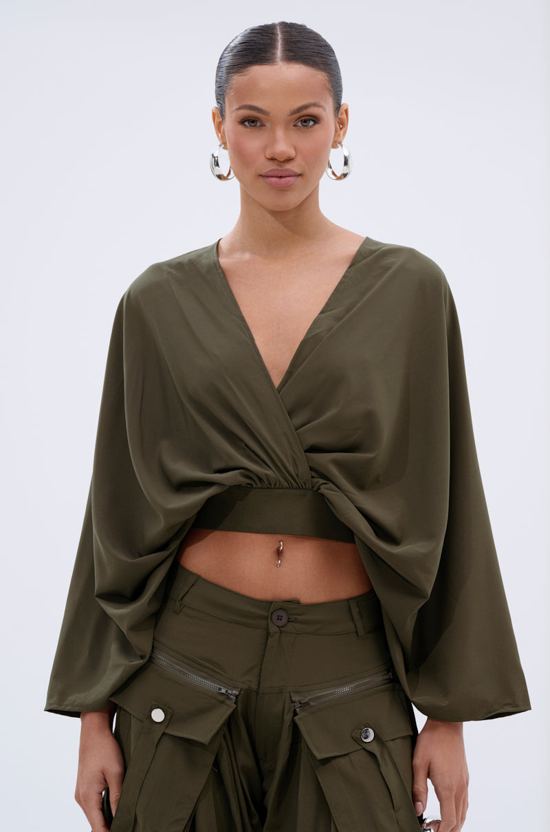 A woman with slicked-back hair wears large hoop earrings and the olive green TALK ABOUT ME BLOUSE by AKIRA Label, featuring a surplice neckline, draped batwing sleeves, and matching cargo pants with zipper and pocket details.