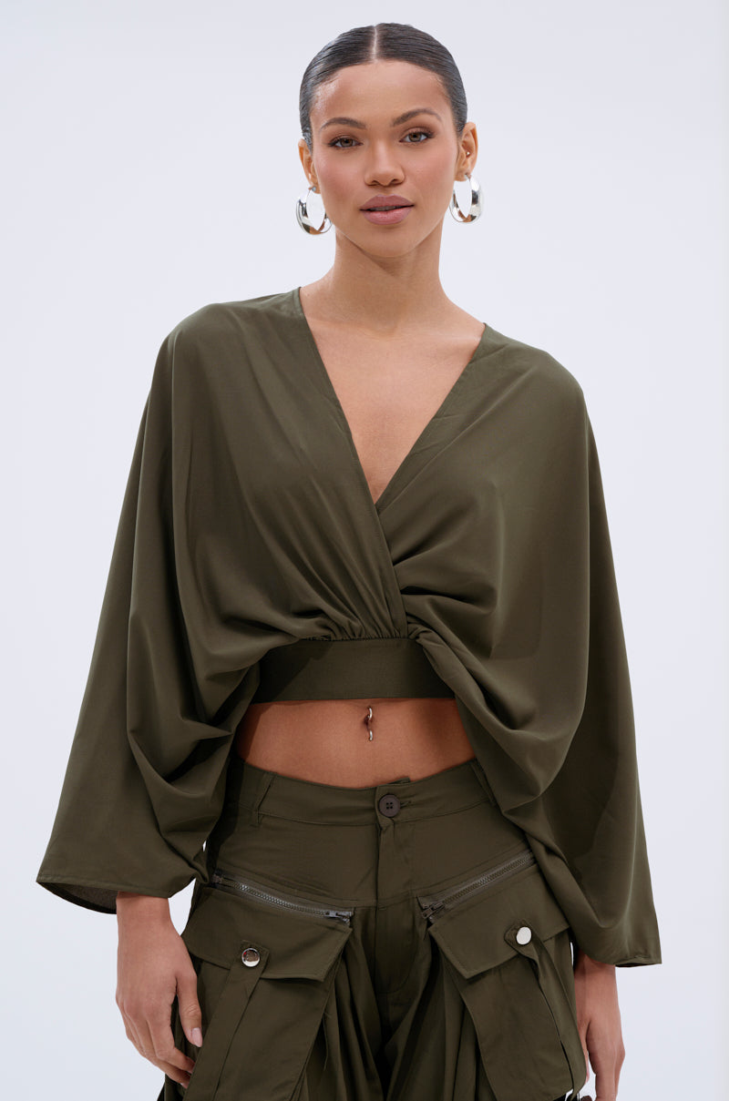 A woman with hoop earrings and slicked-back hair wears the AKIRA Label TALK ABOUT ME BLOUSE, a deep V-neck, flowy olive-green top with batwing sleeves and matching cargo pants, revealing her midriff and navel piercing against a plain light background.