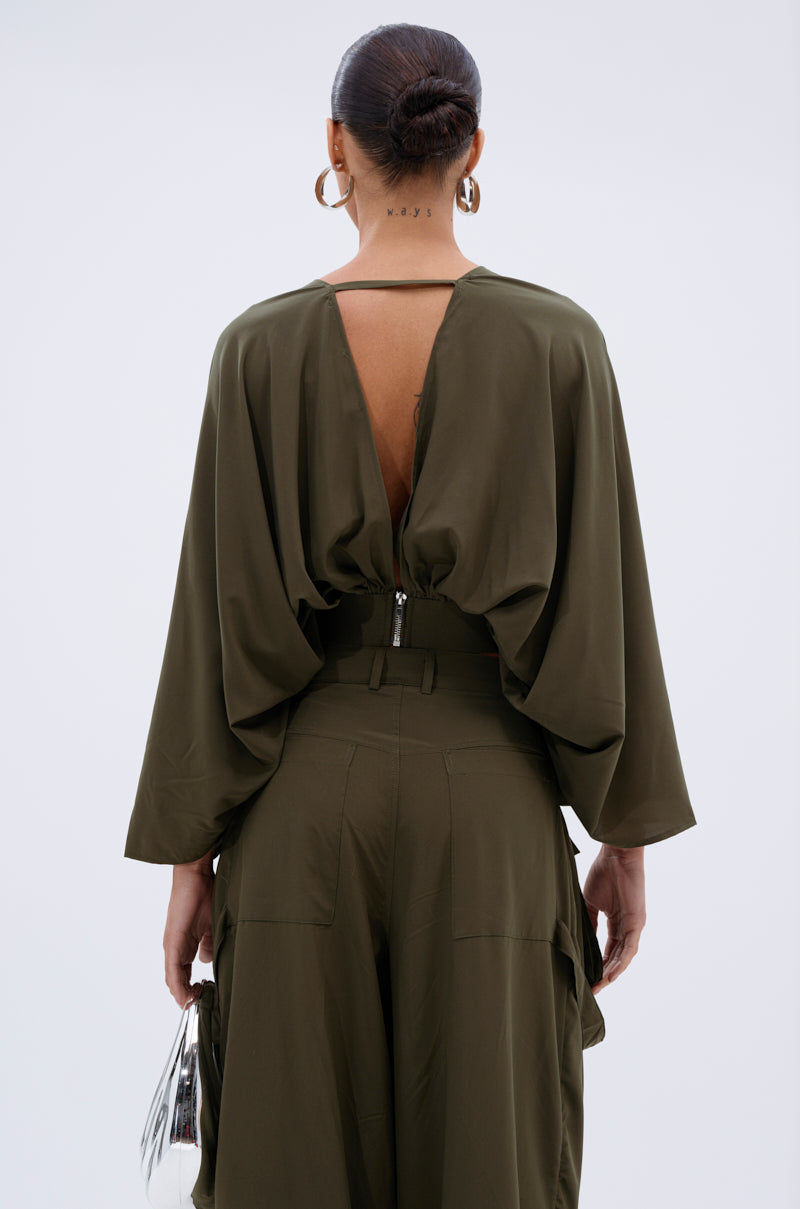 A woman faces away from the camera in an olive-green TALK ABOUT ME BLOUSE with batwing sleeves and an open-back design. Her hair is in a bun, she wears large hoop earrings, and carries a small metallic handbag.