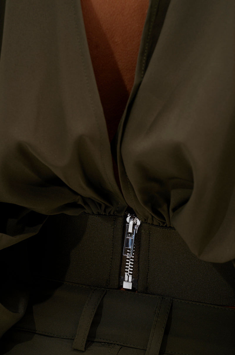 Close-up of an olive green AKIRA Label garment with a silver waist zipper and draped fabric, showing a hint of skin—ideal for pairing with the TALK ABOUT ME BLOUSE.