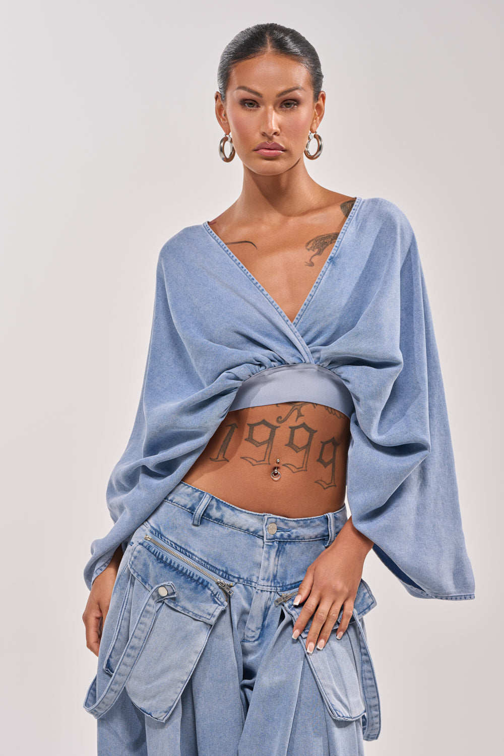 A woman with slicked-back hair and visible tattoos styles the DRIFTING AWAY BLOUSE IN LIGHT BLUE DENIM with baggy blue jeans, large hoop earrings, and creates an elegant day-to-night look. 0