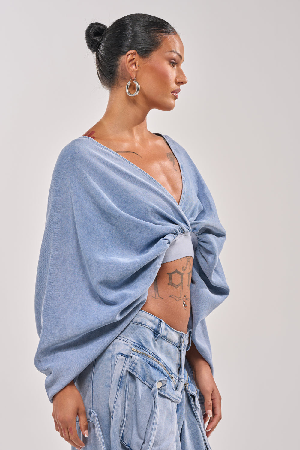A woman with dark hair in a bun wears silver hoop earrings, the DRIFTING AWAY BLOUSE IN LIGHT BLUE DENIM over a white bralette, and loose denim jeans. She has chest and torso tattoos and stands against a plain light background. 2