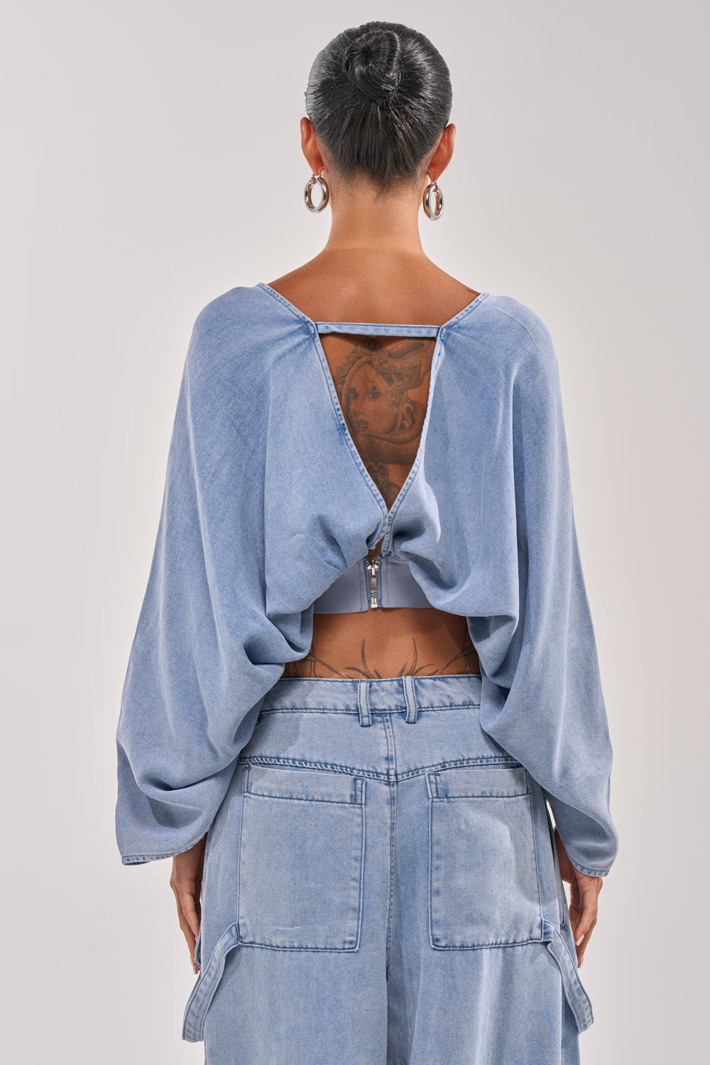 With their hair in a bun and silver hoops, this person wears the DRIFTING AWAY BLOUSE IN LIGHT BLUE DENIM—an open-back style revealing a tattoo—paired with light blue wide-leg jeans, shown from behind. 3