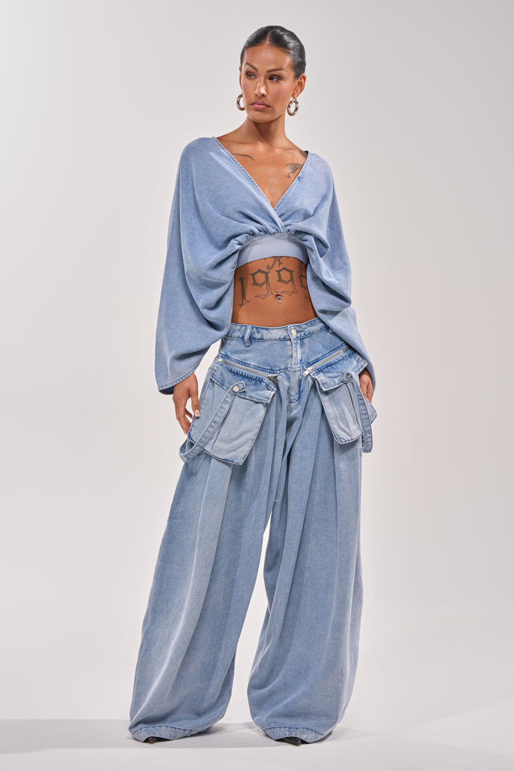 A woman showcases the DRIFTING AWAY BLOUSE IN LIGHT BLUE DENIM, paired with loose cargo denim pants. She accessorizes with hoop earrings, displays torso tattoos, and styles her dark hair back for a chic day-to-night look. 4