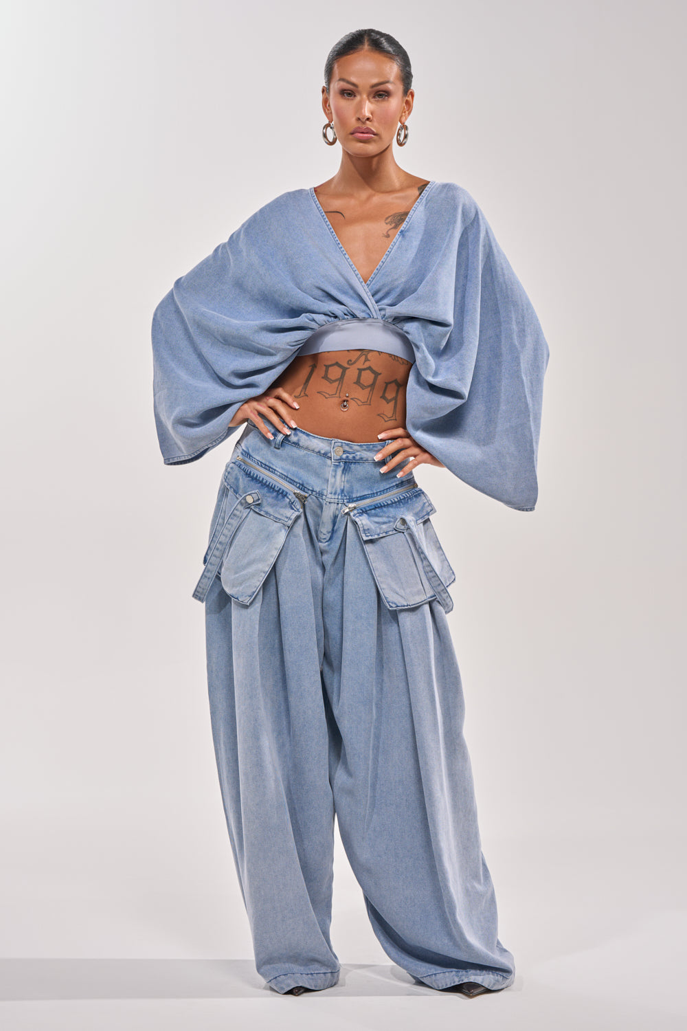 A woman stands confidently in the DRIFTING AWAY BLOUSE IN LIGHT BLUE DENIM and oversized pants, with a "1999" tattoo on her midriff and hoop earrings accenting her look—perfect for effortless day-to-night style. 5