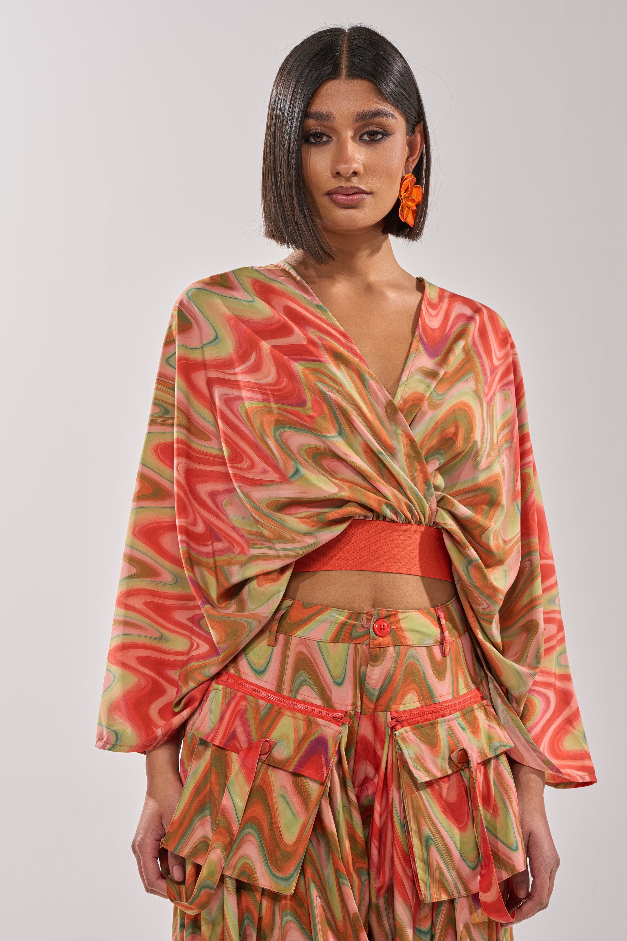 A woman models festival style in the DRIFTING AWAY BLOUSE, an abstract swirl wrap top with wide sleeves, paired with matching high-waisted pants featuring large pockets. She finishes the cropped look with straight hair and orange statement earrings. 0