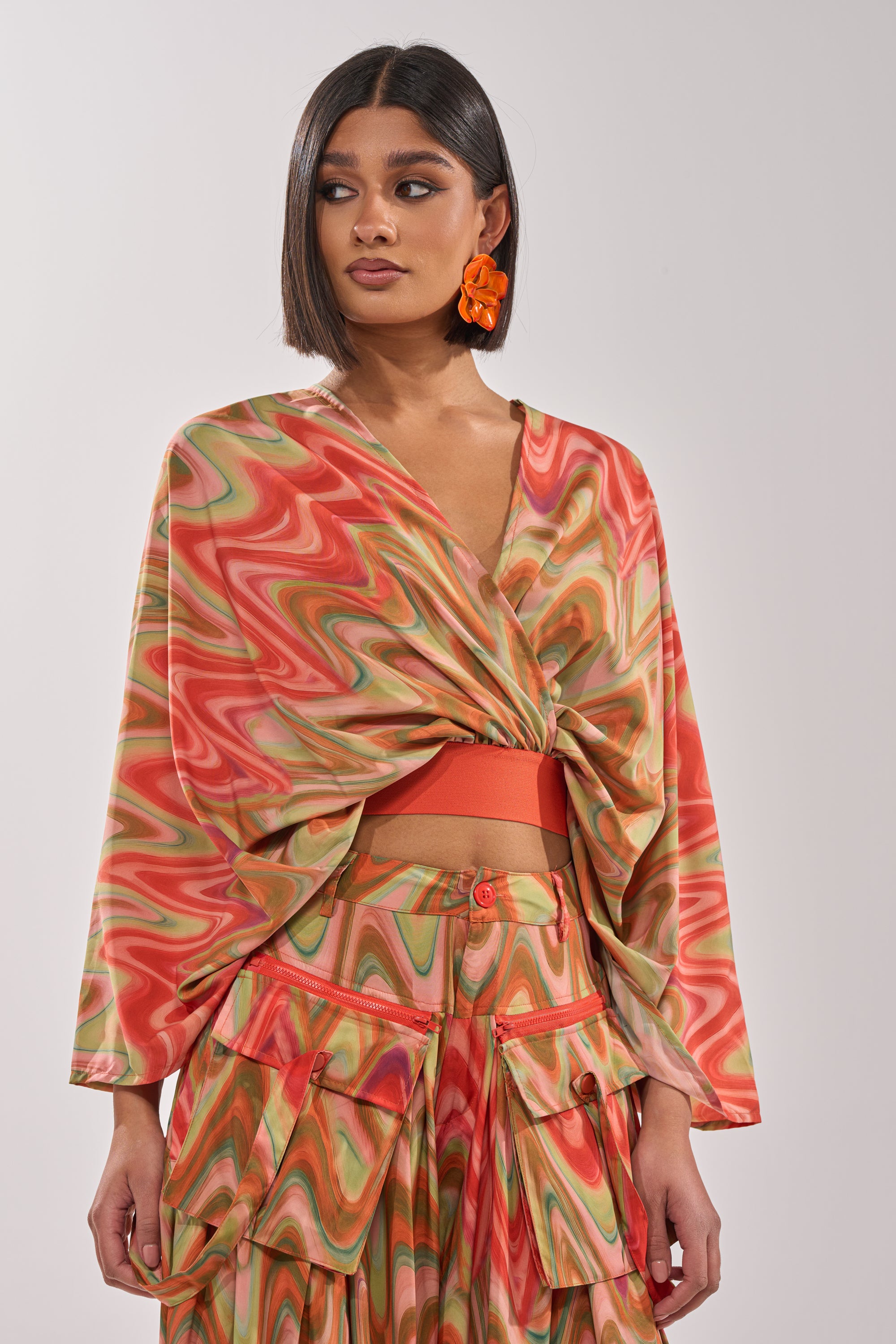 A woman with a sleek bob wears the DRIFTING AWAY BLOUSE—a cropped, abstract swirl wrap top in red, orange, and green—paired with bold orange flower earrings. The playful look stands out against a plain, light background. 1