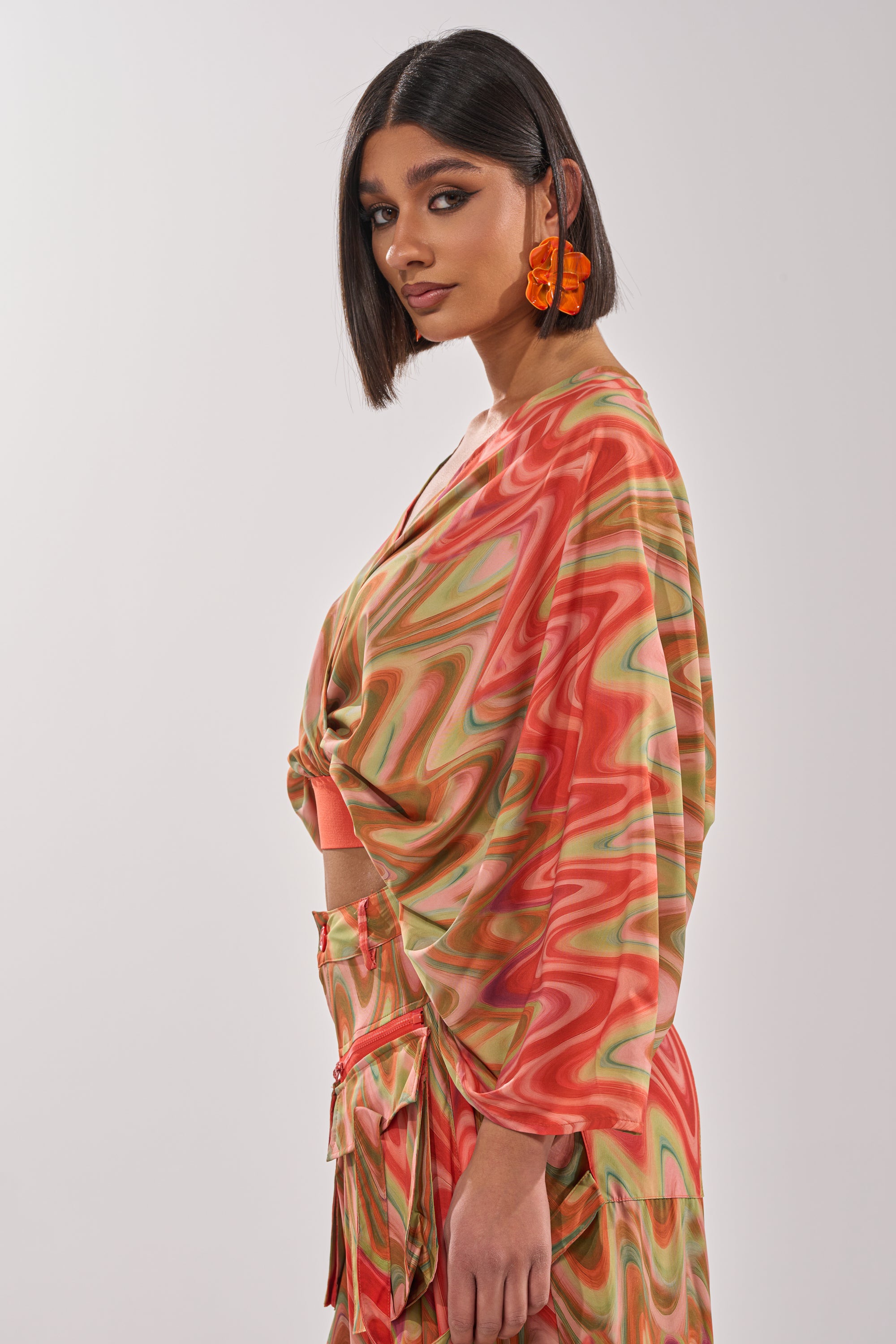 A woman with a sleek bob wears orange statement earrings and the DRIFTING AWAY BLOUSE—sunset-toned with colorful wavy patterns, loose sleeves, and a knotted front—standing against a simple light background. 2