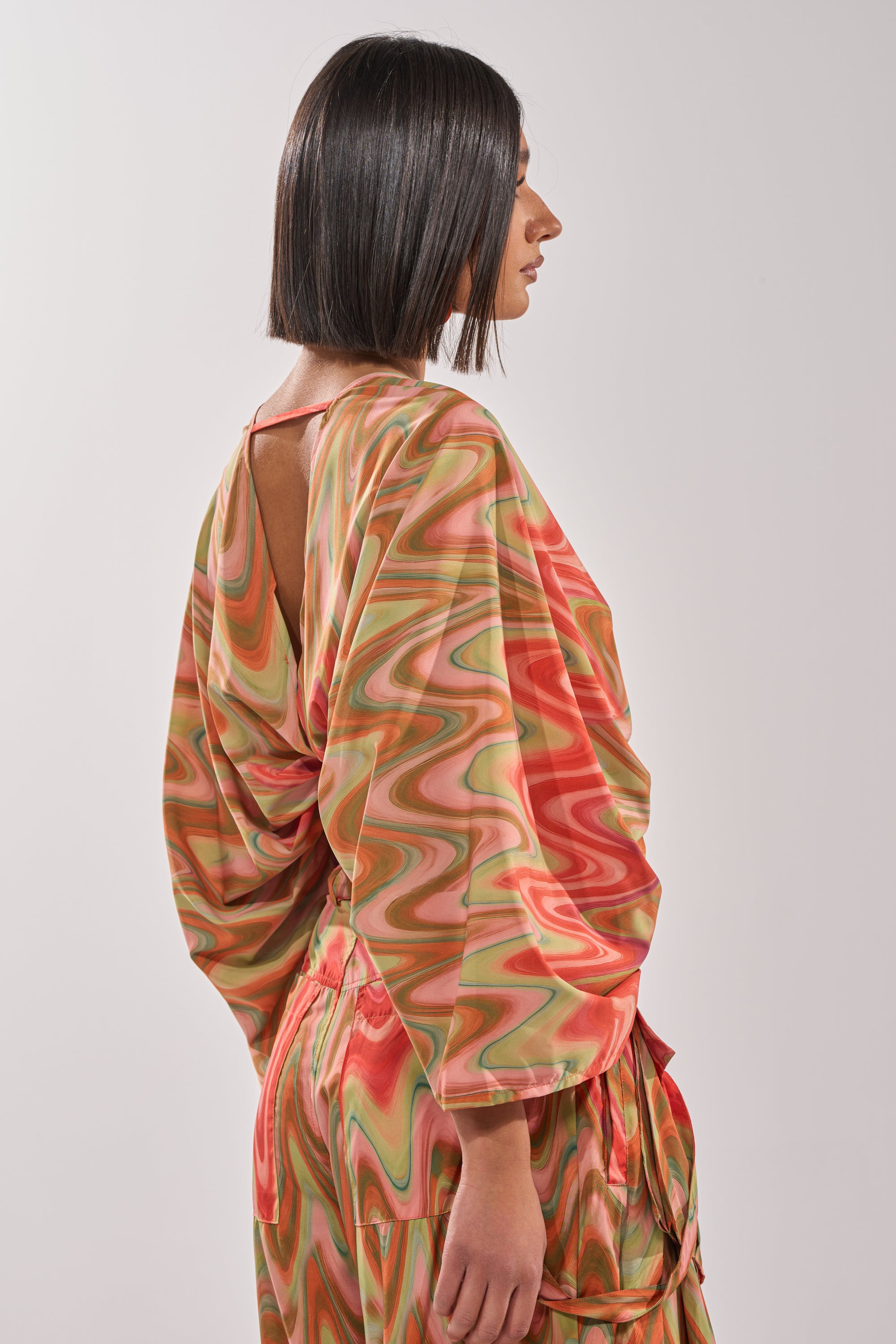 Wearing the DRIFTING AWAY BLOUSE—a loose, long-sleeved wrap top with abstract orange, green, and pink swirls—a person with a sleek bob stands in profile against a plain light background. 3