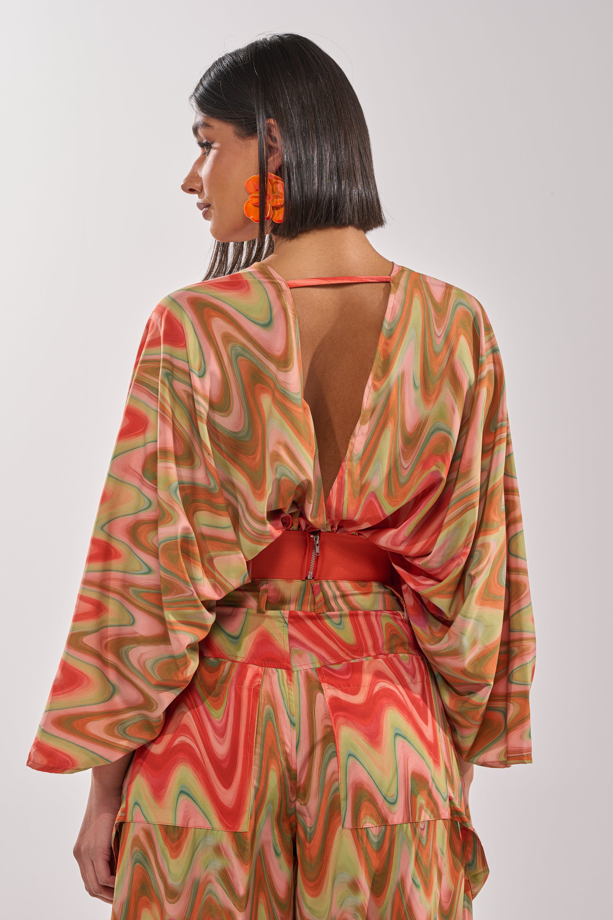 A woman with short dark hair and orange earrings faces away, showcasing the bold, festival style and open-back details of the DRIFTING AWAY BLOUSE—a cropped wrap top with a colorful abstract swirl print. 4