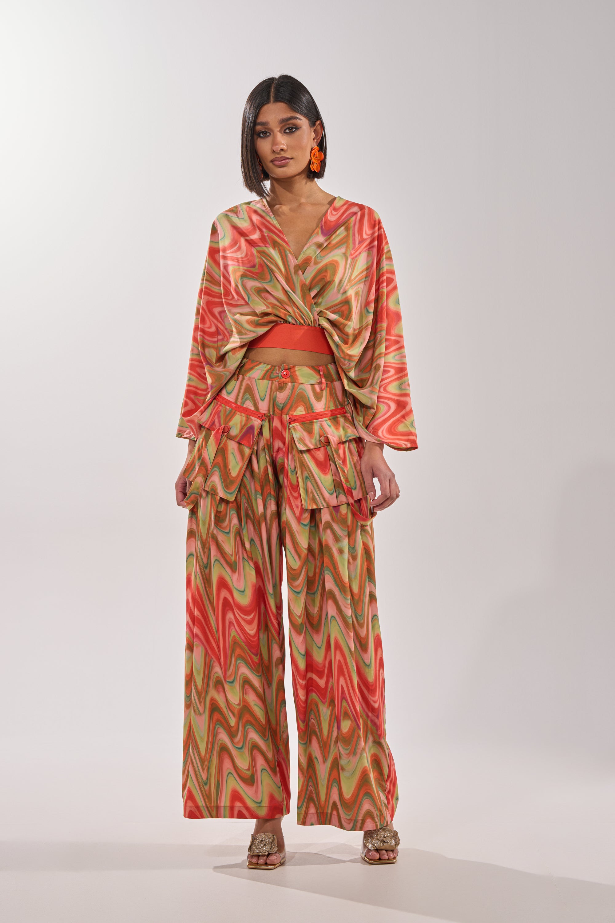 A woman with bobbed hair, orange earrings, and open-toed shoes poses in the DRIFTING AWAY BLOUSE—an abstract swirl wrap top—in sunset hues with wide-leg pants, facing forward against a plain background. 5