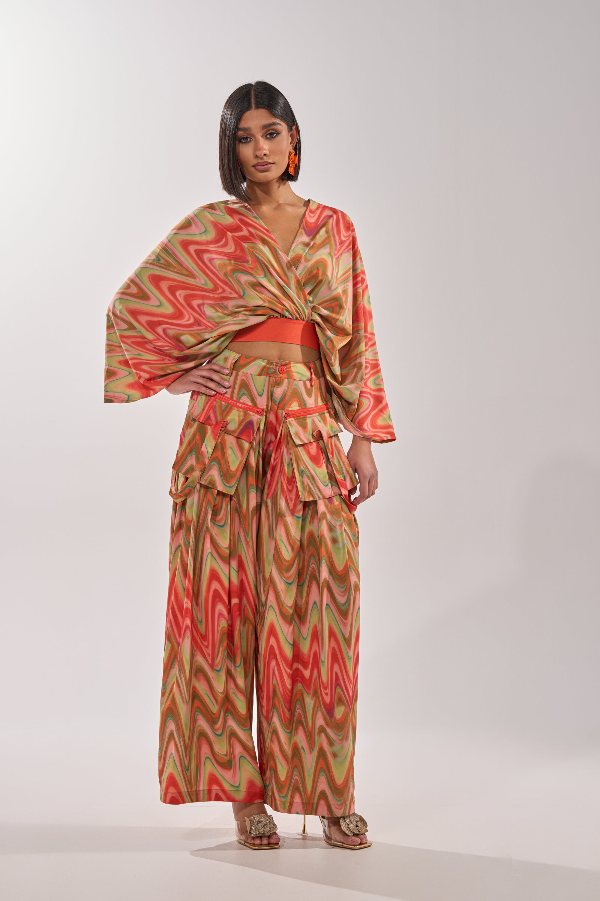 A woman stands wearing the DRIFTING AWAY BLOUSE, a colorful abstract swirl wrap top with wide sleeves and sunset tones, paired with wide-legged pants, gold sandals, shoulder-length straight hair, and large orange earrings. 7