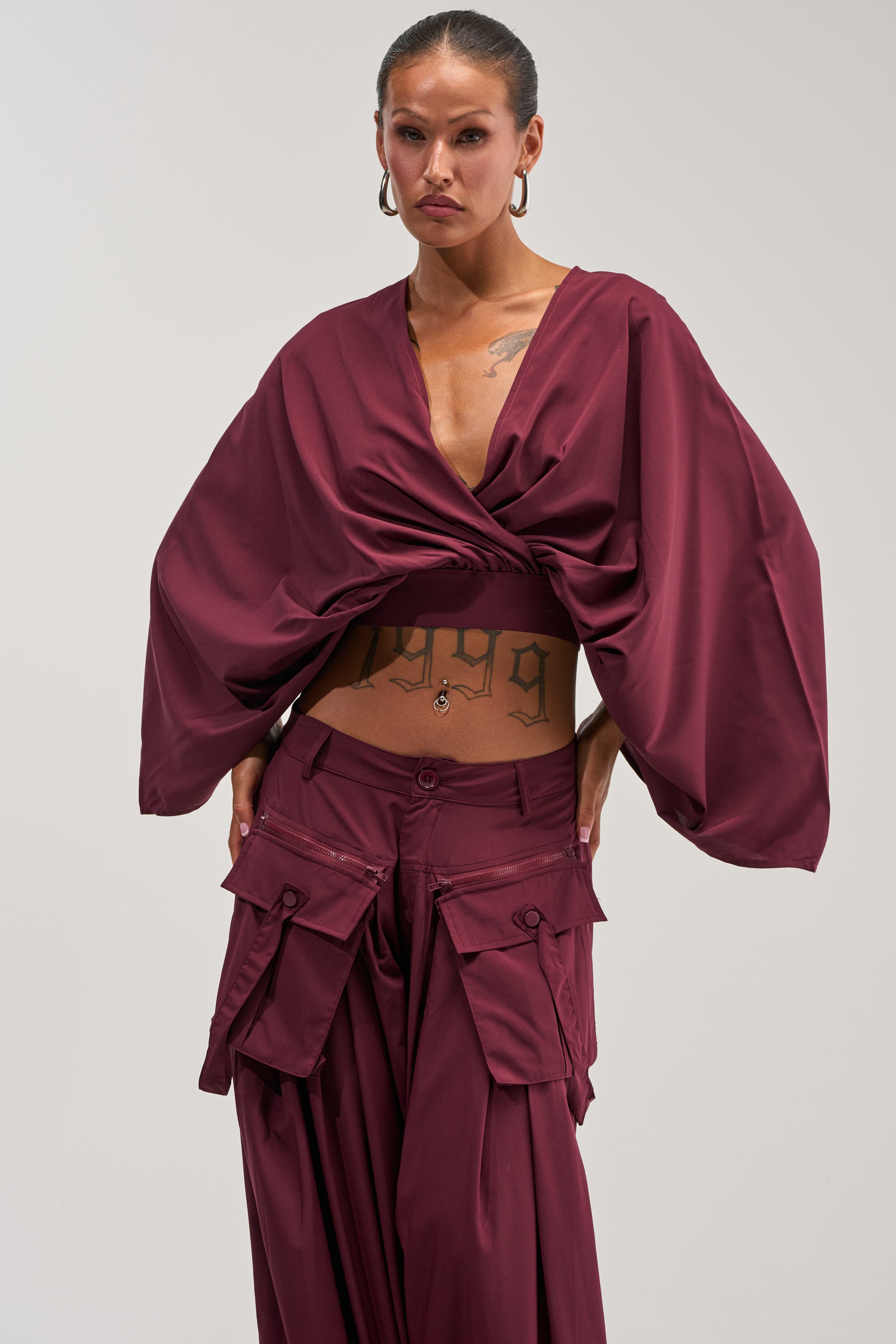 A person stands confidently against a plain light background, showcasing the TALK ABOUT ME BLOUSE IN DEEP BURGUNDY—a bold top featuring wide sleeves and cargo pockets—while visible tattoos on their torso add a modern edge to the stylish office look.