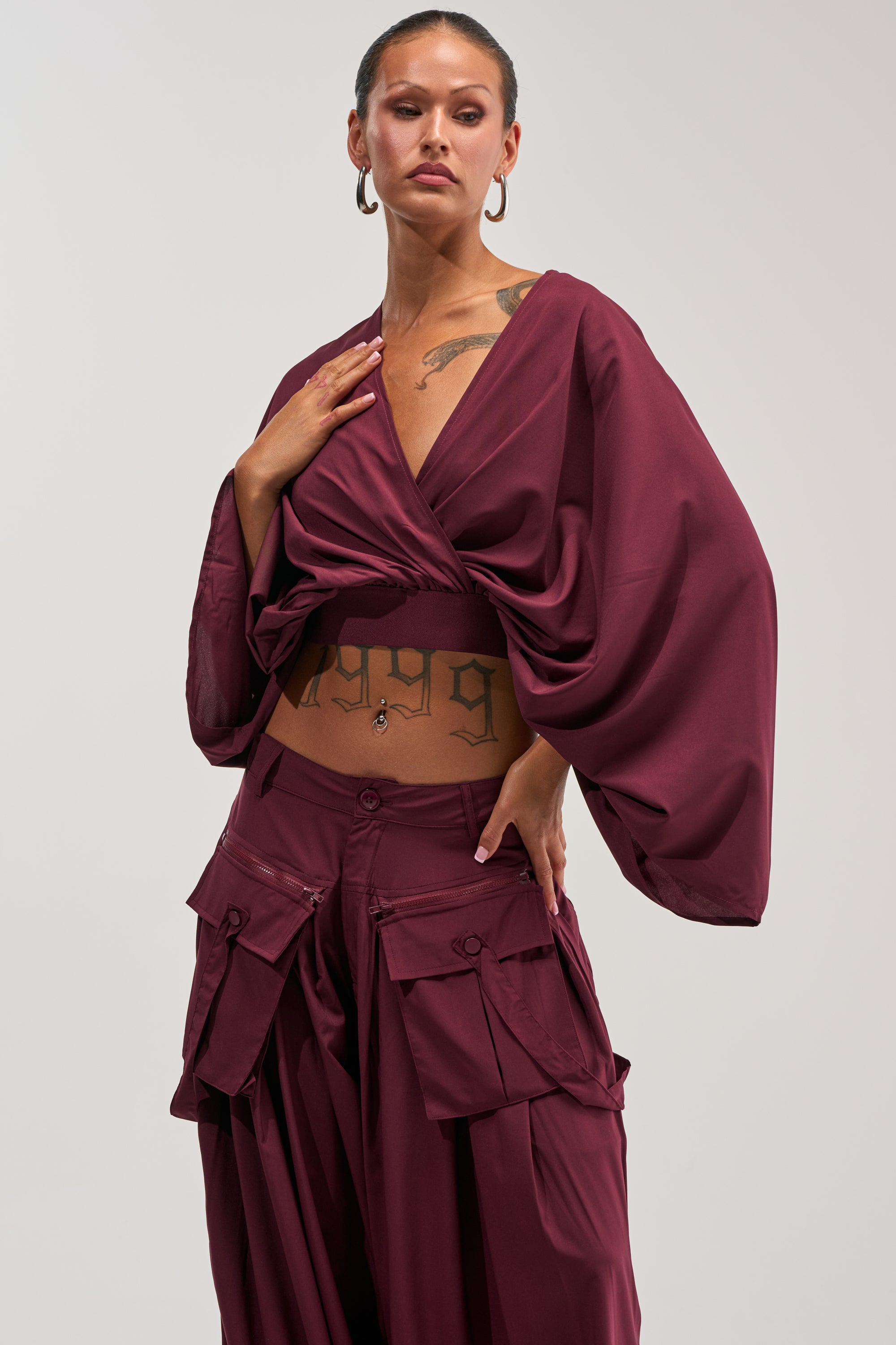 Wearing the TALK ABOUT ME BLOUSE IN DEEP BURGUNDY with flowy sleeves and paired with cargo pants, this individual exudes confidence. They accessorize with hoop earrings and display tattoos on their chest and stomach, while neutral makeup completes the look—a bold interpretation of office style that fuses personal flair with sophistication.