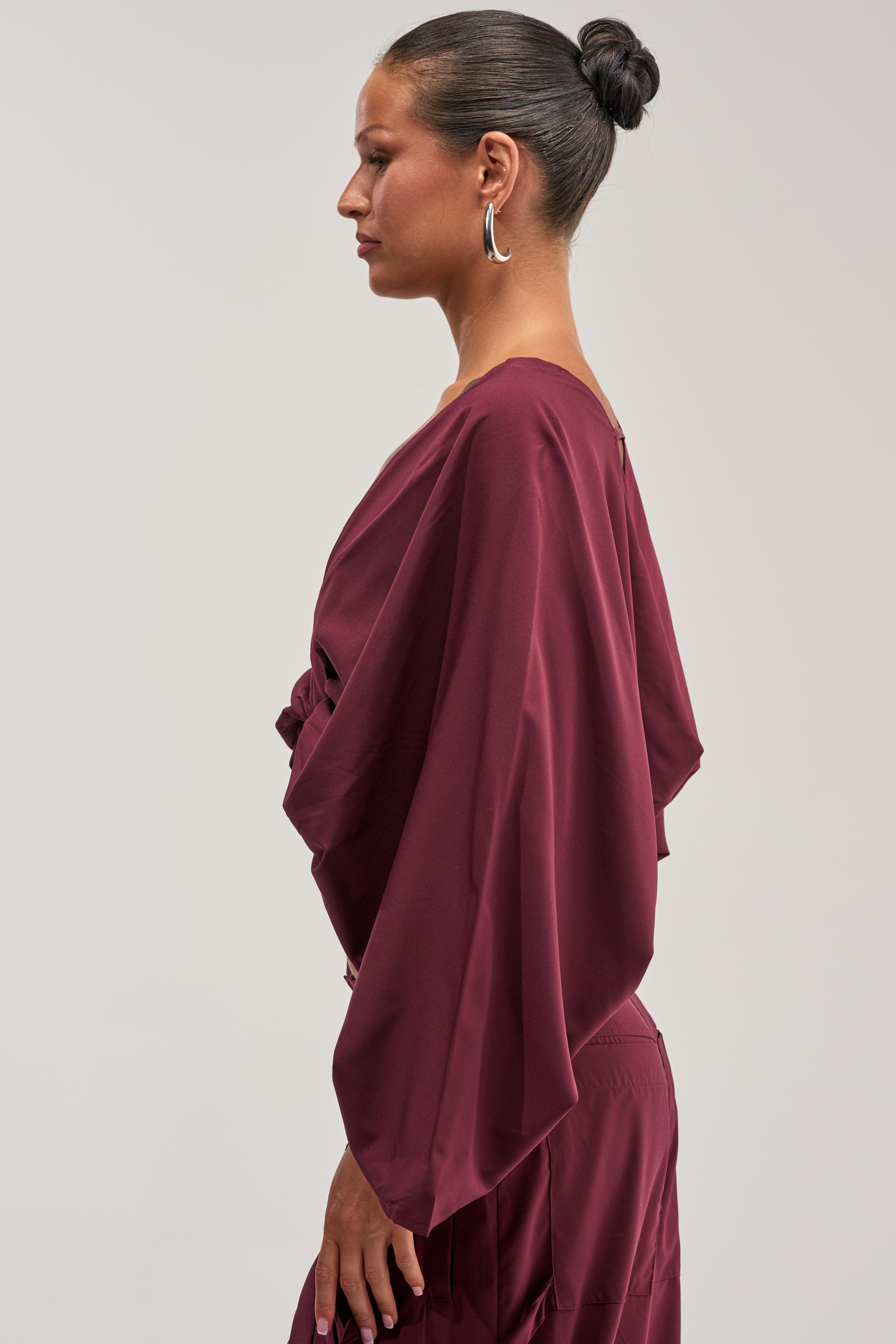 A woman with dark hair in a bun wears the TALK ABOUT ME BLOUSE IN DEEP BURGUNDY, featuring wide sleeves and styled with large hoop earrings, as she stands in profile against a plain light background—making an elegant choice for modern office styling.