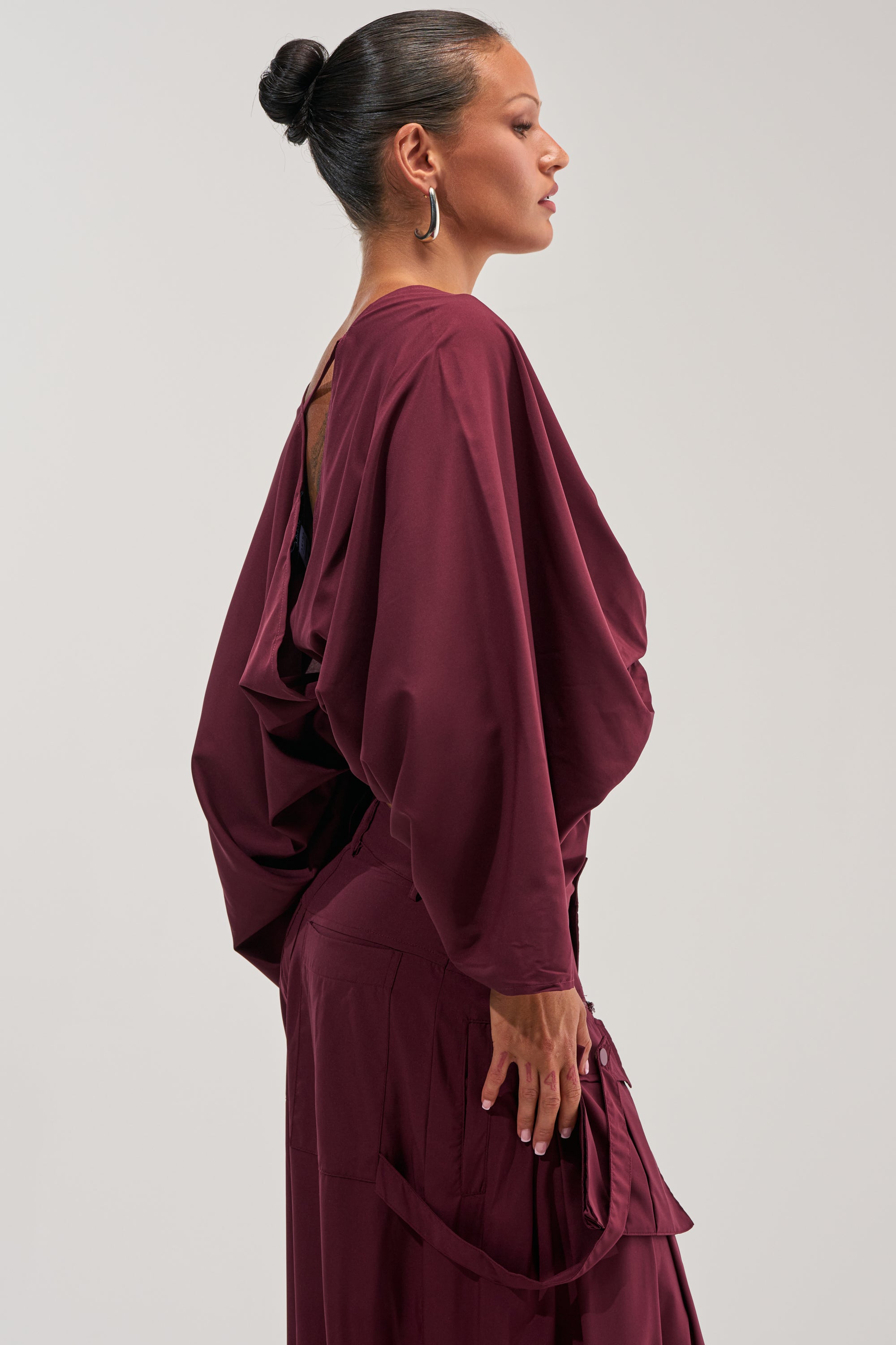 A woman in profile with her hair styled in a bun wears the TALK ABOUT ME BLOUSE IN DEEP BURGUNDY, featuring flowing draped sleeves, standing against a plain light background—an elegant choice for office styling.