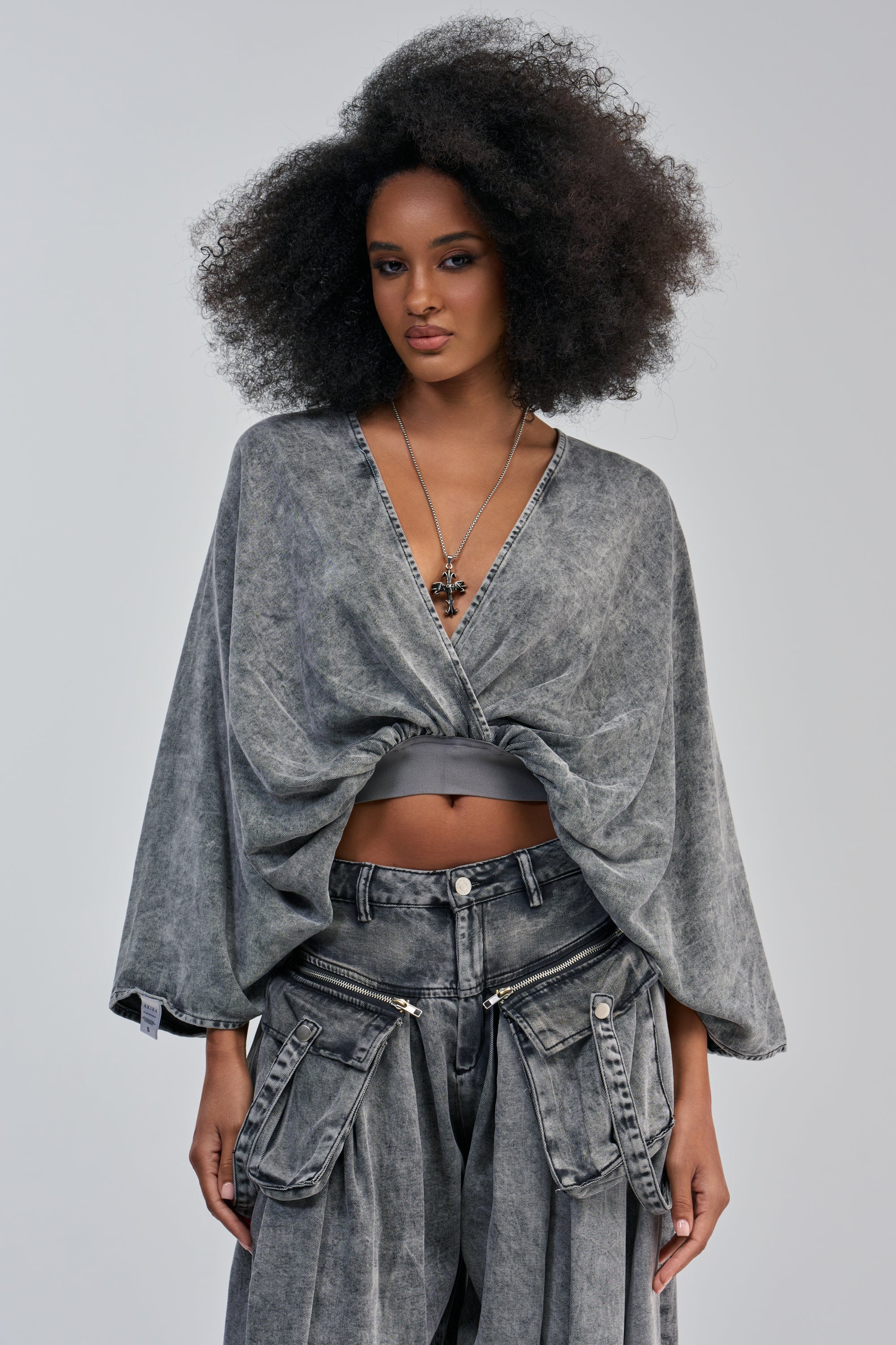 A model with a voluminous afro poses in a grey acid-wash denim outfit. She wears the DRIFTING AWAY BLOUSE IN CHARCOAL, a draped, kimono-sleeve V-neck top featuring an open-back design, styled with a grey bandeau and matching cargo-style jeans with silver zippered pockets and a cross pendant necklace. This statement streetwear look is set against a light grey background.
