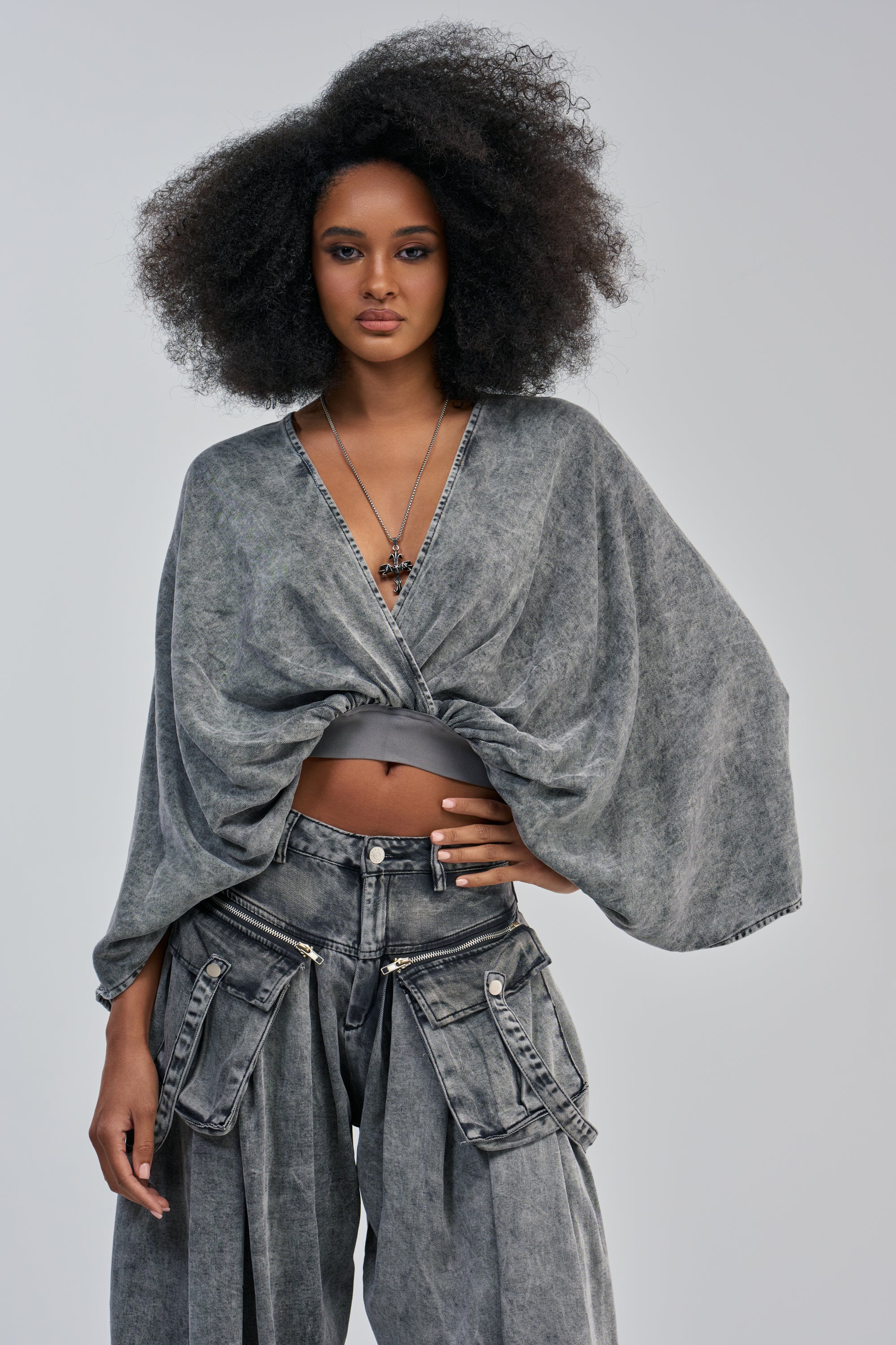 A woman with voluminous curly hair showcases a bold streetwear style in the DRIFTING AWAY BLOUSE IN CHARCOAL, featuring a deep V-neck and wide sleeves, paired with matching baggy charcoal denim pants with large side pockets. She poses confidently against a plain light background, gazing directly at the camera.