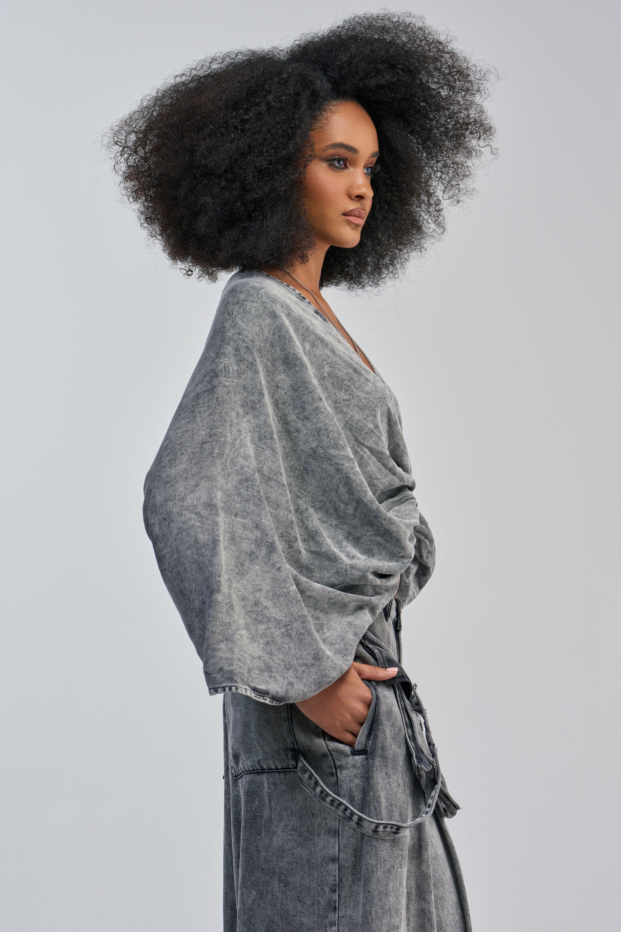 A model with a voluminous afro hairstyle stands in profile against a grey background, wearing the DRIFTING AWAY BLOUSE IN CHARCOAL with oversized dolman sleeves and matching pants, her hand casually tucked into her pocket for a distinct streetwear vibe.