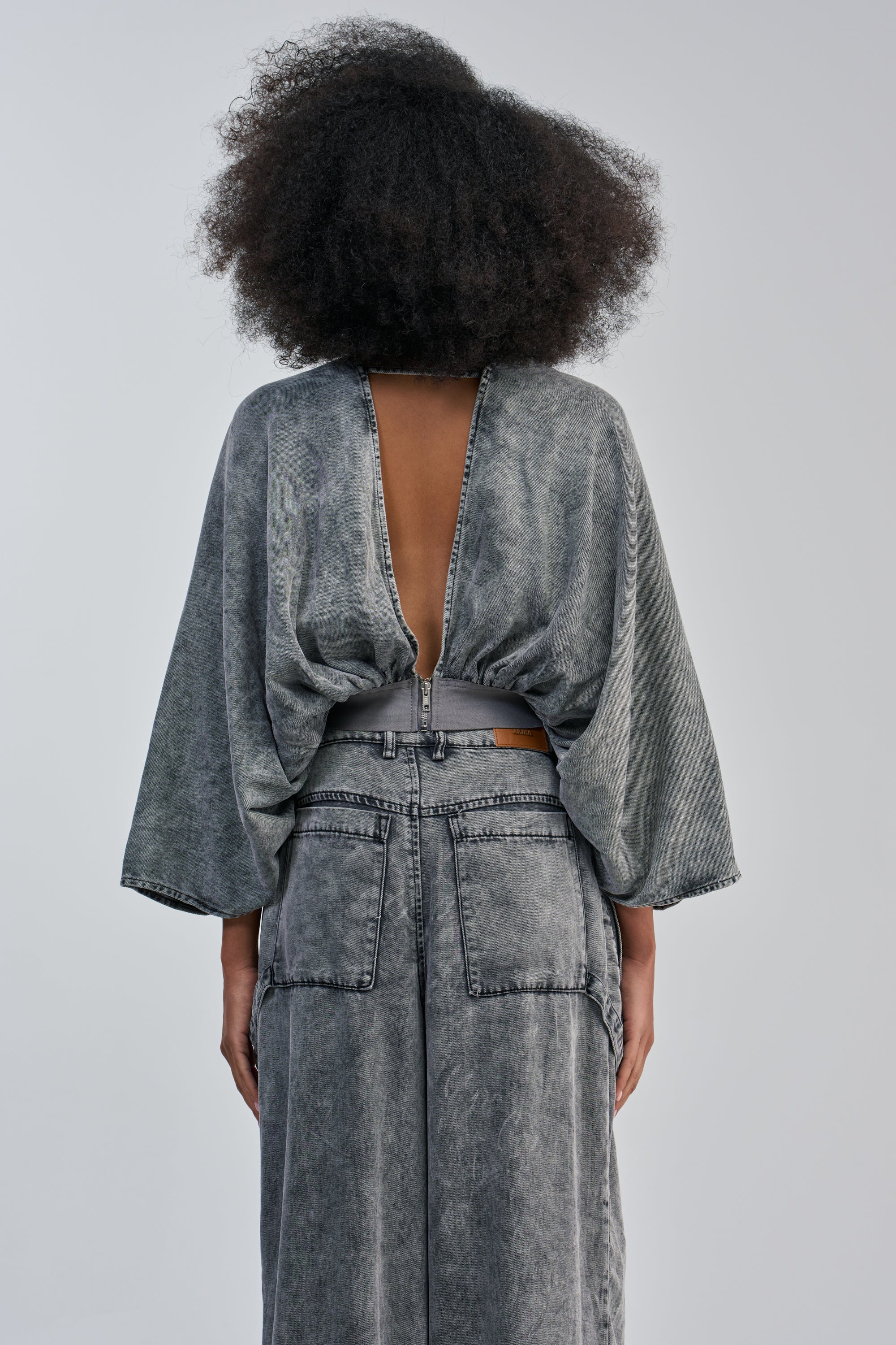 Wearing the DRIFTING AWAY BLOUSE IN CHARCOAL, a person with curly hair showcases an effortlessly chic streetwear look with its loose fit, oversized dolman sleeves, and open back as they stand against a plain light background.