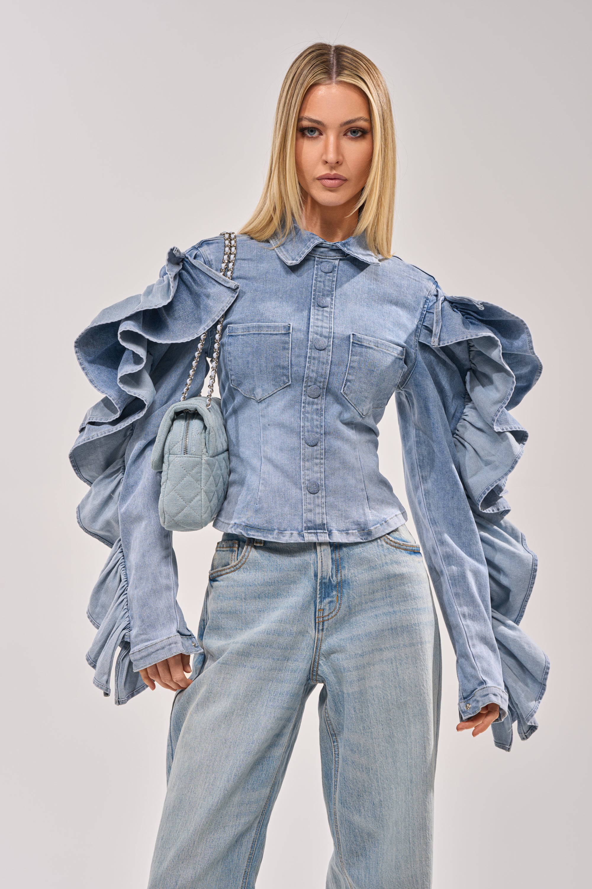 A woman models the DREA RUFFLE SLEEVE DENIM BUTTON DOWN with matching light blue jeans and a small quilted handbag. She has straight blonde hair and poses confidently against a plain background. 0