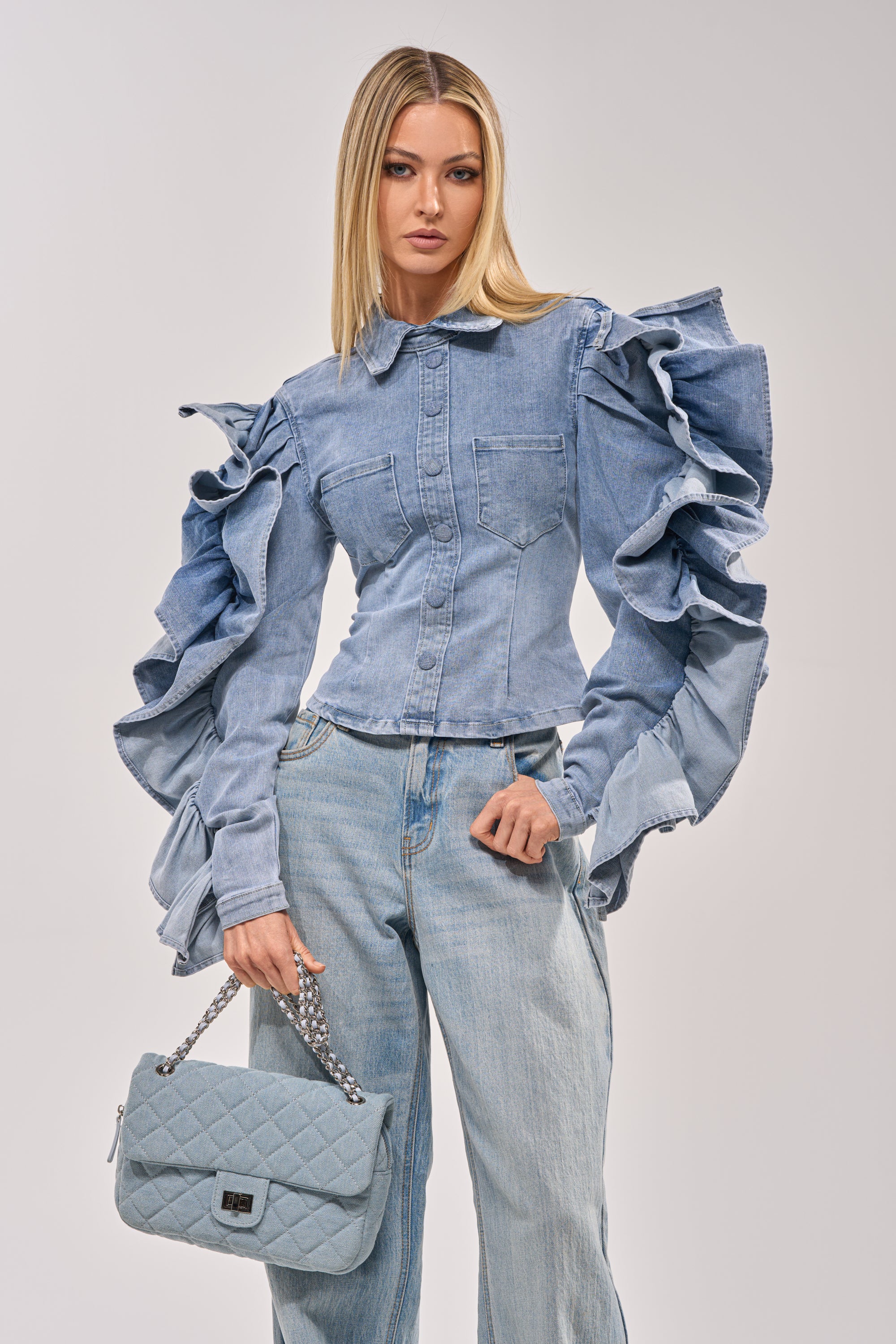 Wearing the DREA RUFFLE SLEEVE DENIM BUTTON DOWN with light blue jeans and a quilted handbag, a woman creates a bold brunch look against a simple backdrop. 1